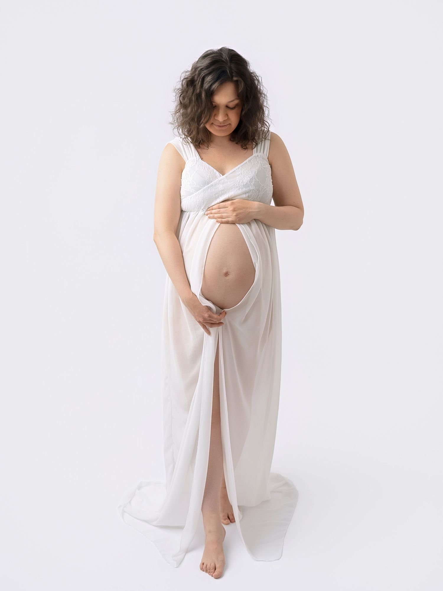 Pregnant woman in white flowing maternity gown cradling baby bump against white background.