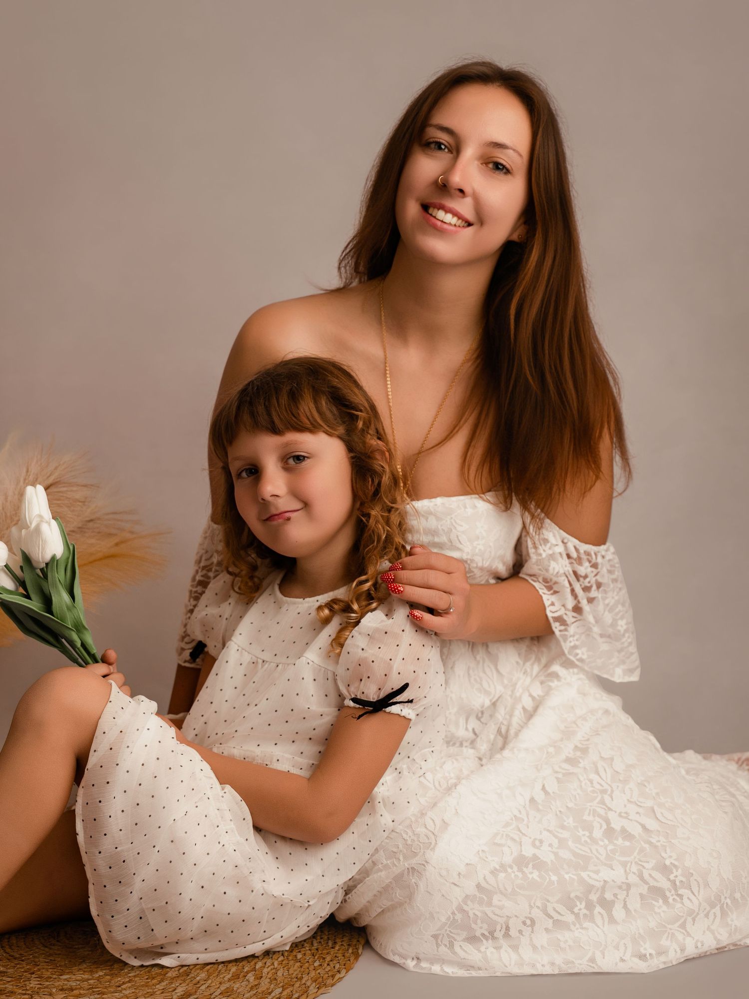A woman and young girl in matching white dresses pose together, with the girl holding white tulips.
