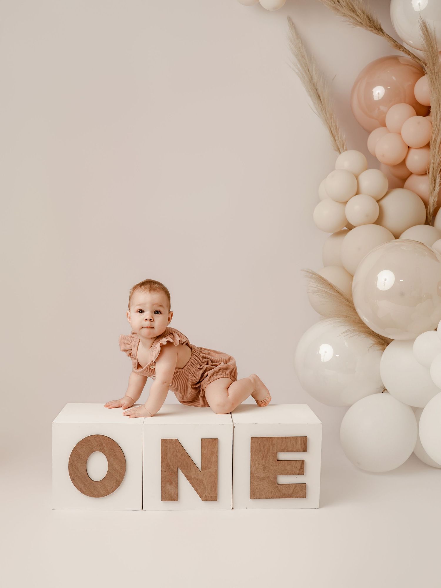 A baby in a brown romper poses on white ONE letter blocks surrounded by neutral balloons for a first birthday photo.