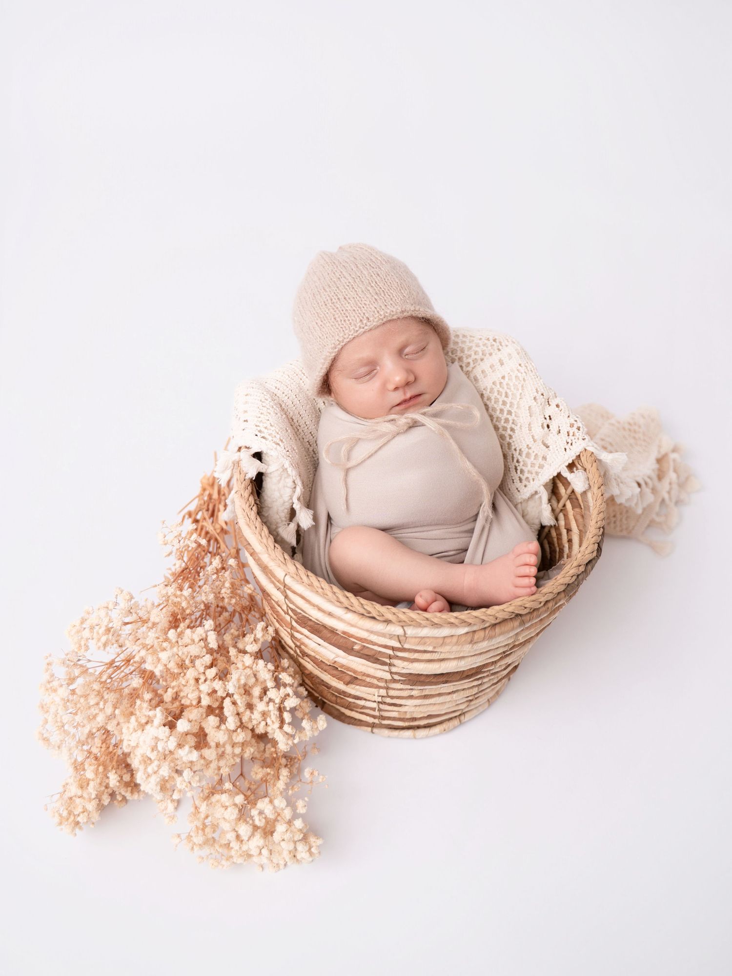 Peaceful newborn baby sleeping in a wicker basket with beige knit hat and dried flowers.