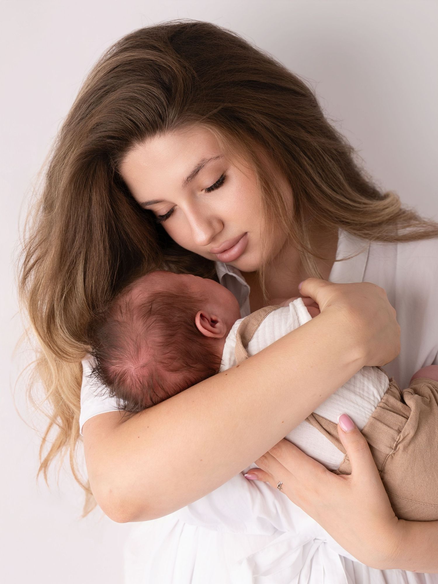 Young mother with long brown hair tenderly holds sleeping newborn baby close to her chest in soft white light.
