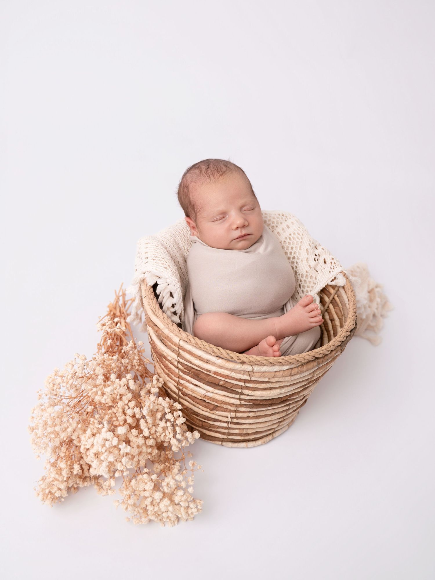 Sleeping newborn baby in a wicker basket with cream knit blanket and dried flowers on white background.