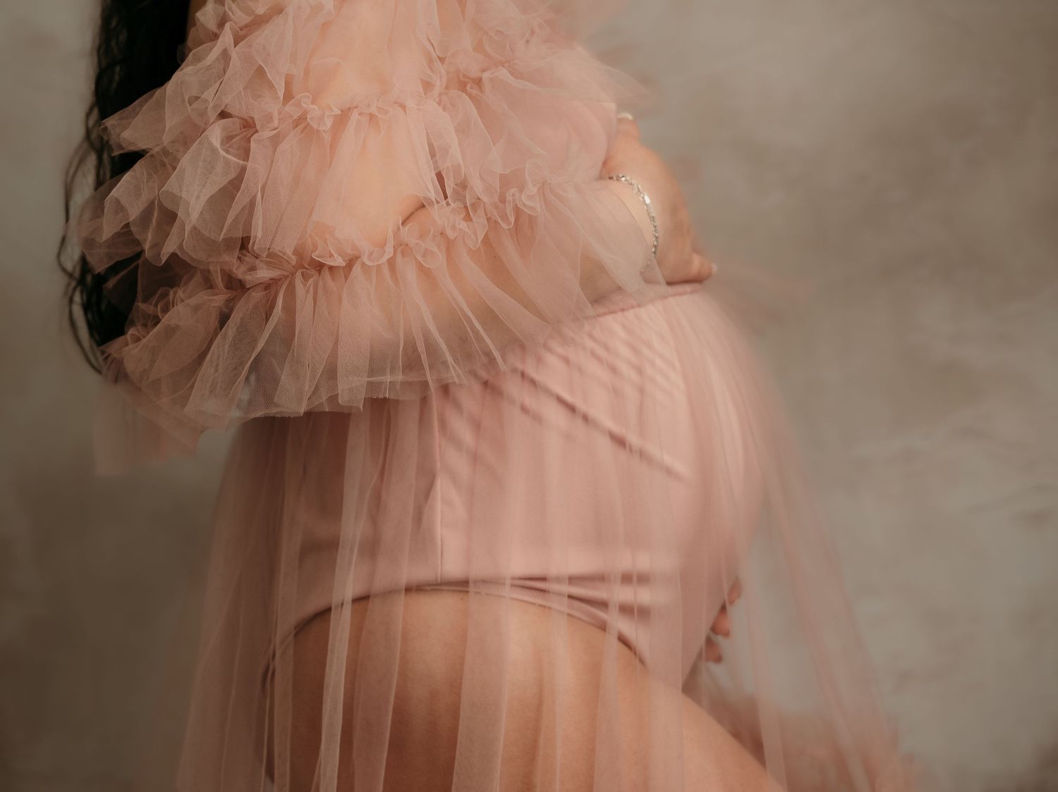 Close-up of a pregnant woman in a sheer blush pink ruffled tulle maternity gown highlighting her baby bump.
