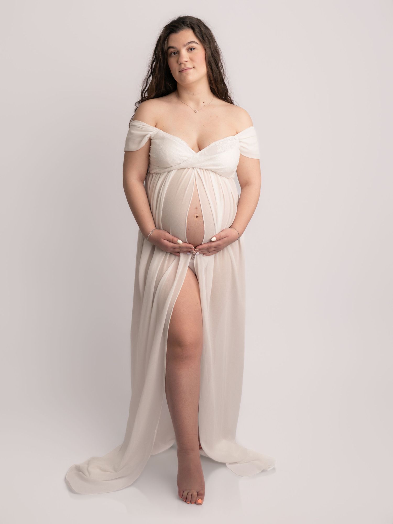 Pregnant woman in sheer white off-shoulder maternity gown with front slit cradling bare baby bump in studio.