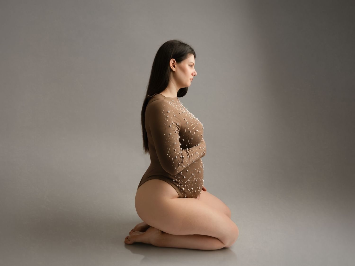 Woman in pearl-studded brown bodysuit kneeling gracefully on studio floor, side profile.