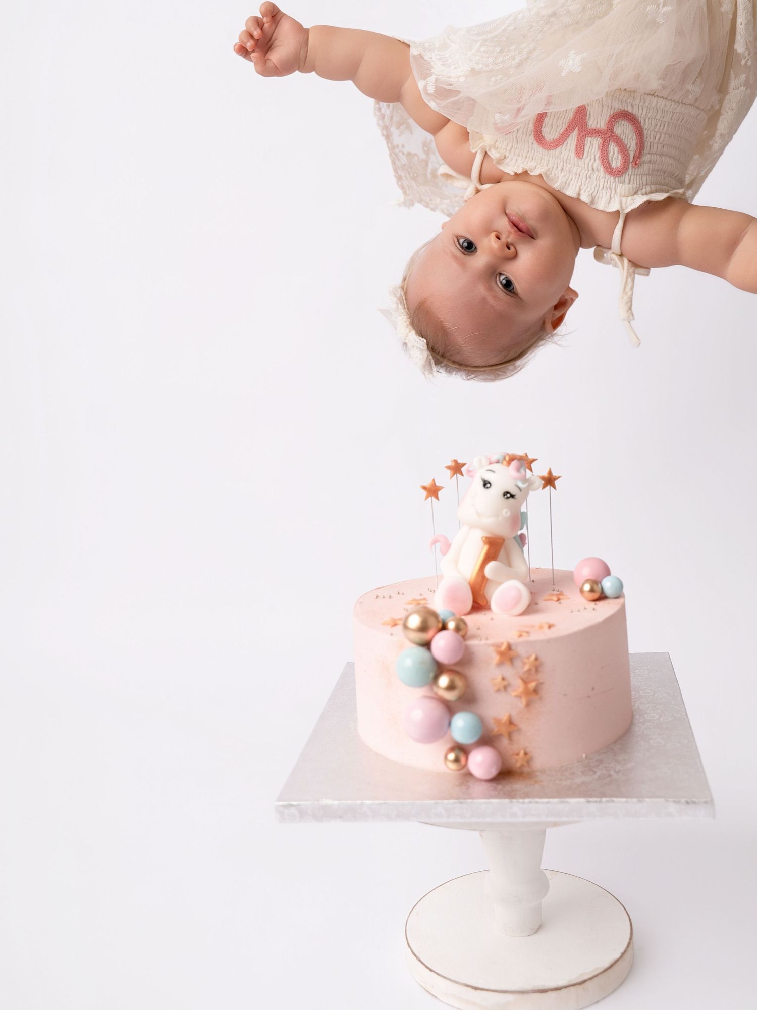 Baby girl in white dress dangles upside down above a pink unicorn first birthday cake with pastel balloon decorations.