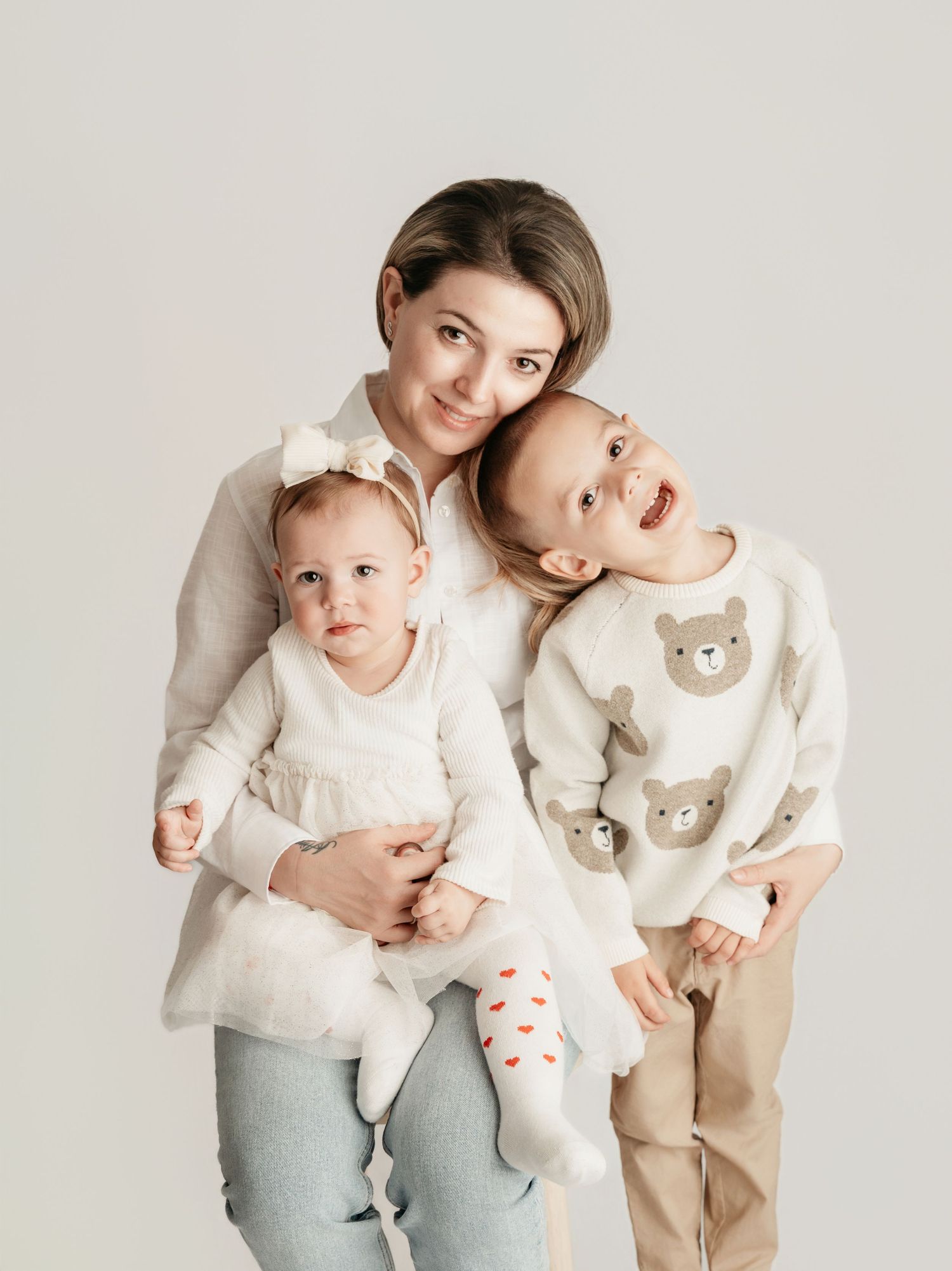 A smiling mother holds her baby girl and young son in matching bear outfits in a soft studio portrait.