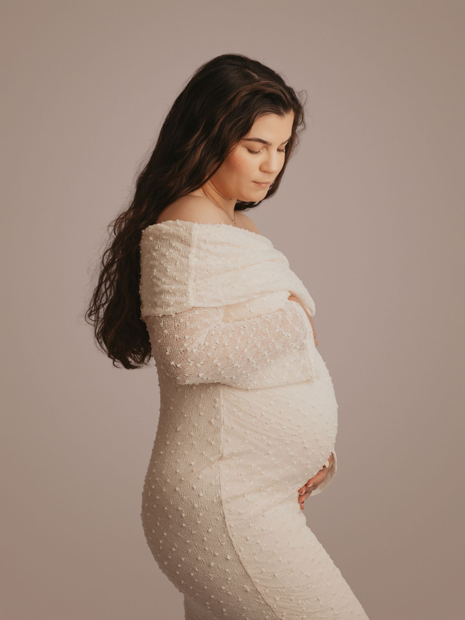 Pregnant woman in white off-shoulder lace maternity gown looking down, cradling baby bump in studio.