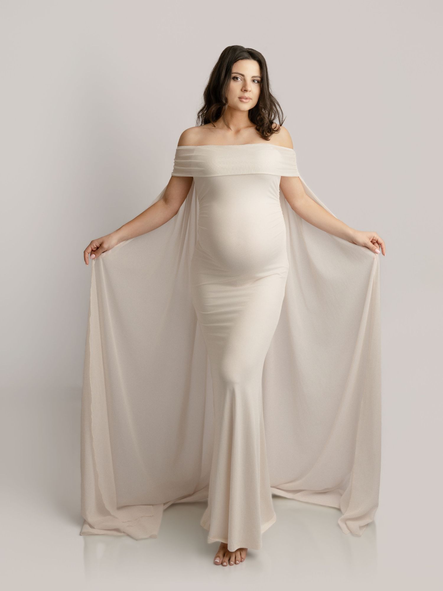 Pregnant woman in elegant off-shoulder cream gown with flowing cape sleeves poses gracefully in studio.