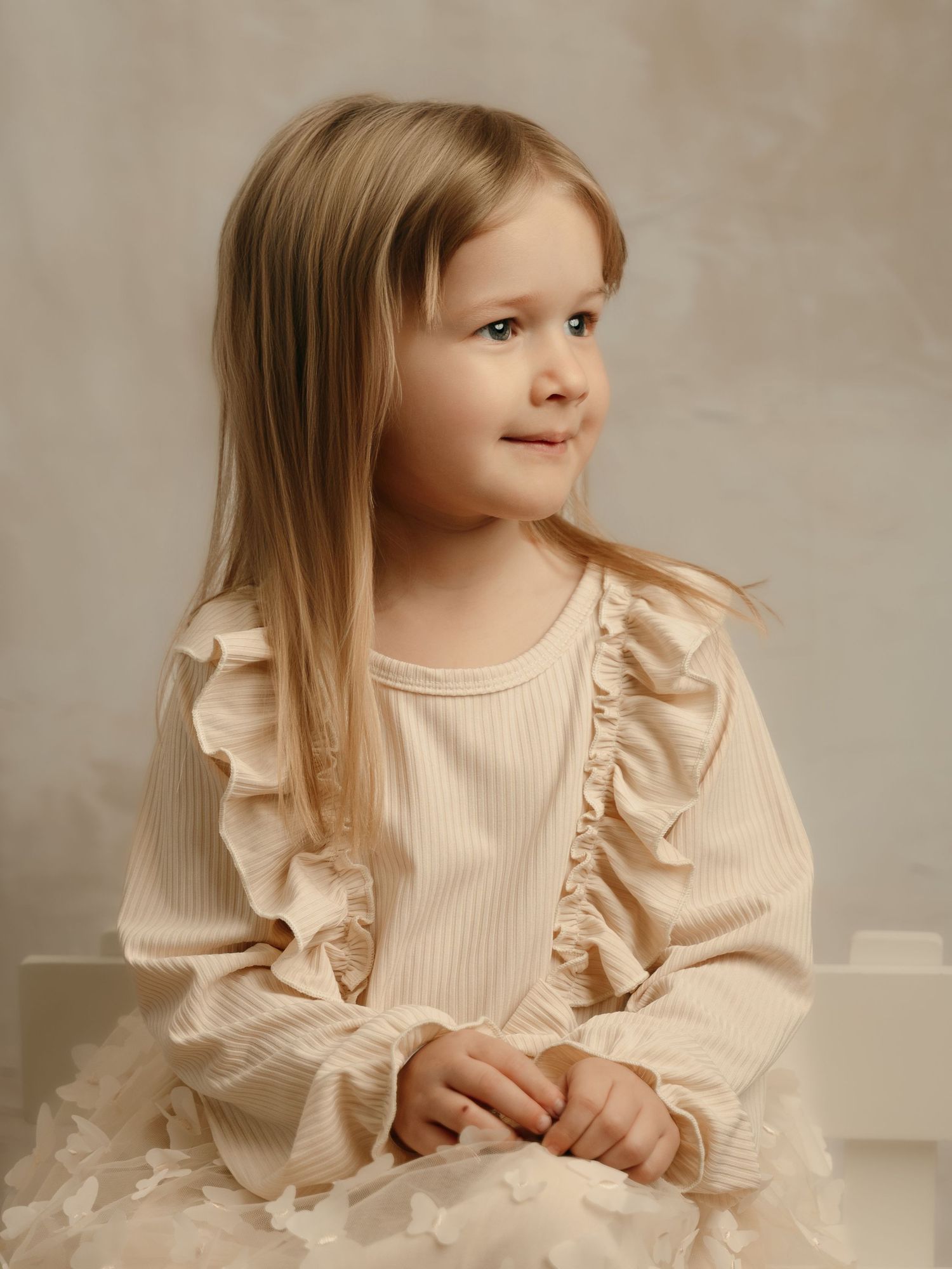 Young girl with blonde hair wearing a cream ribbed ruffle dress, sitting gracefully and looking to the side.