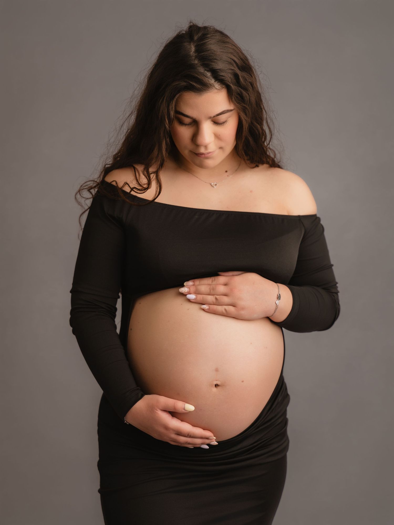 Pregnant woman in black off-shoulder maternity dress looking down while gently holding bare baby bump.