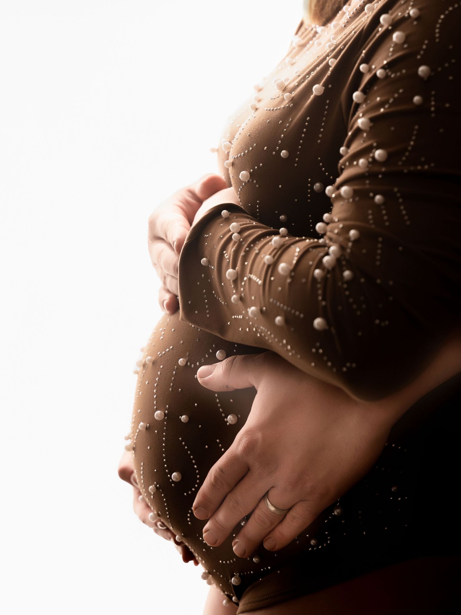 Pregnant woman in brown pearl-embellished bodysuit cradling baby bump, side profile maternity photo.