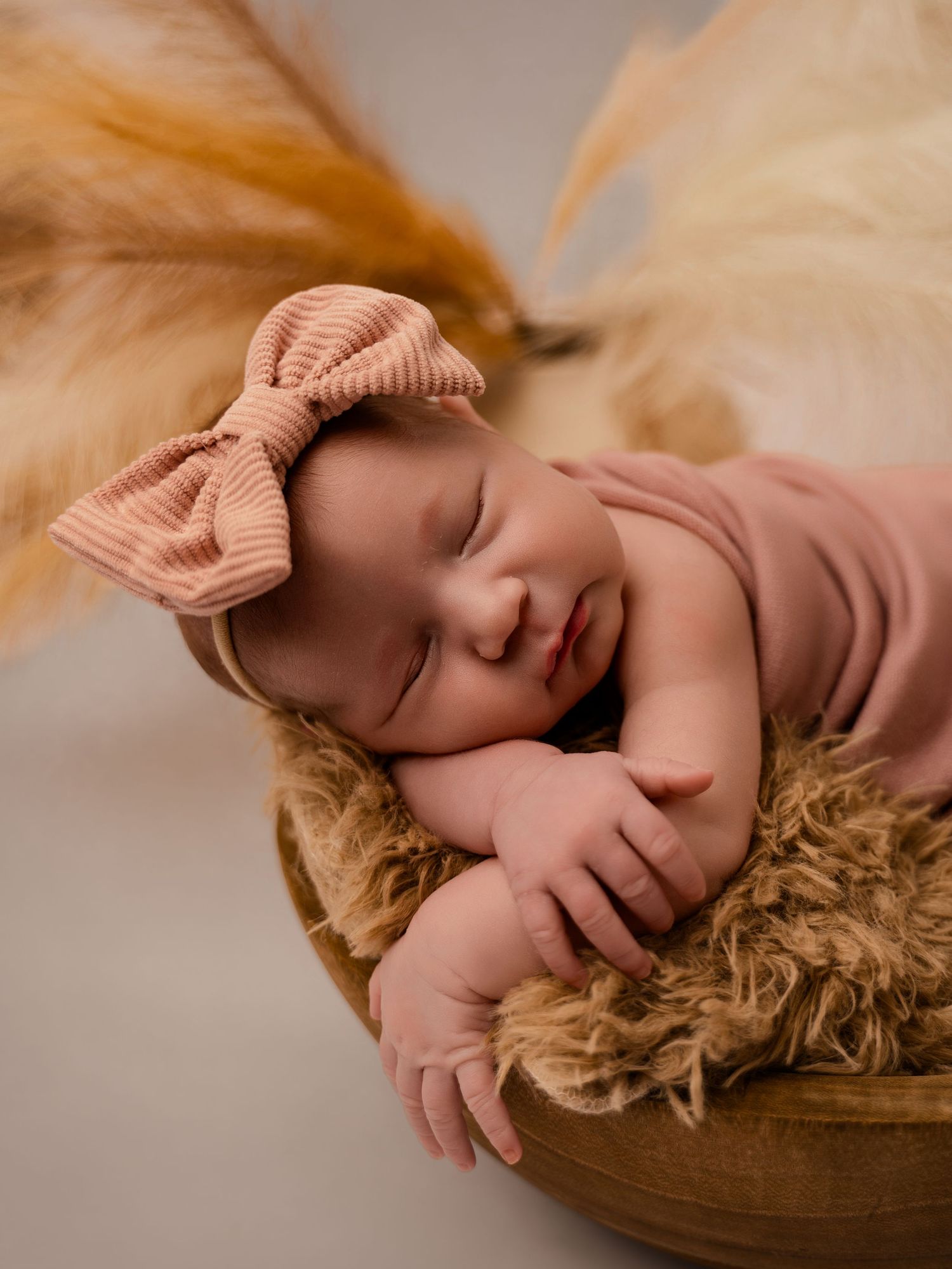 Peaceful newborn baby girl in pink bow headband sleeping in wooden bowl with fluffy fur lining.