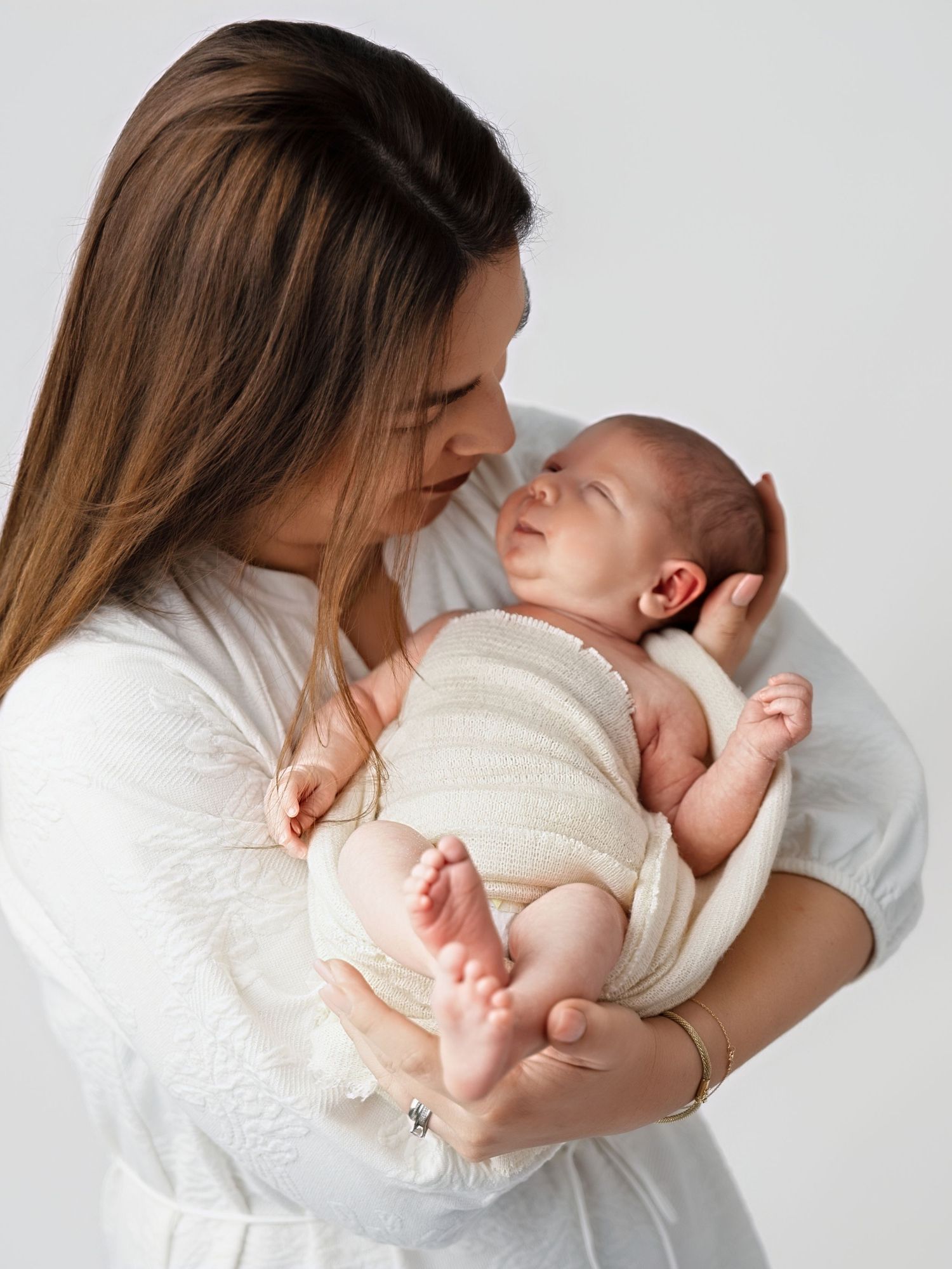 A mother in white gently cradles her newborn baby, sharing a tender moment against a soft gray background.