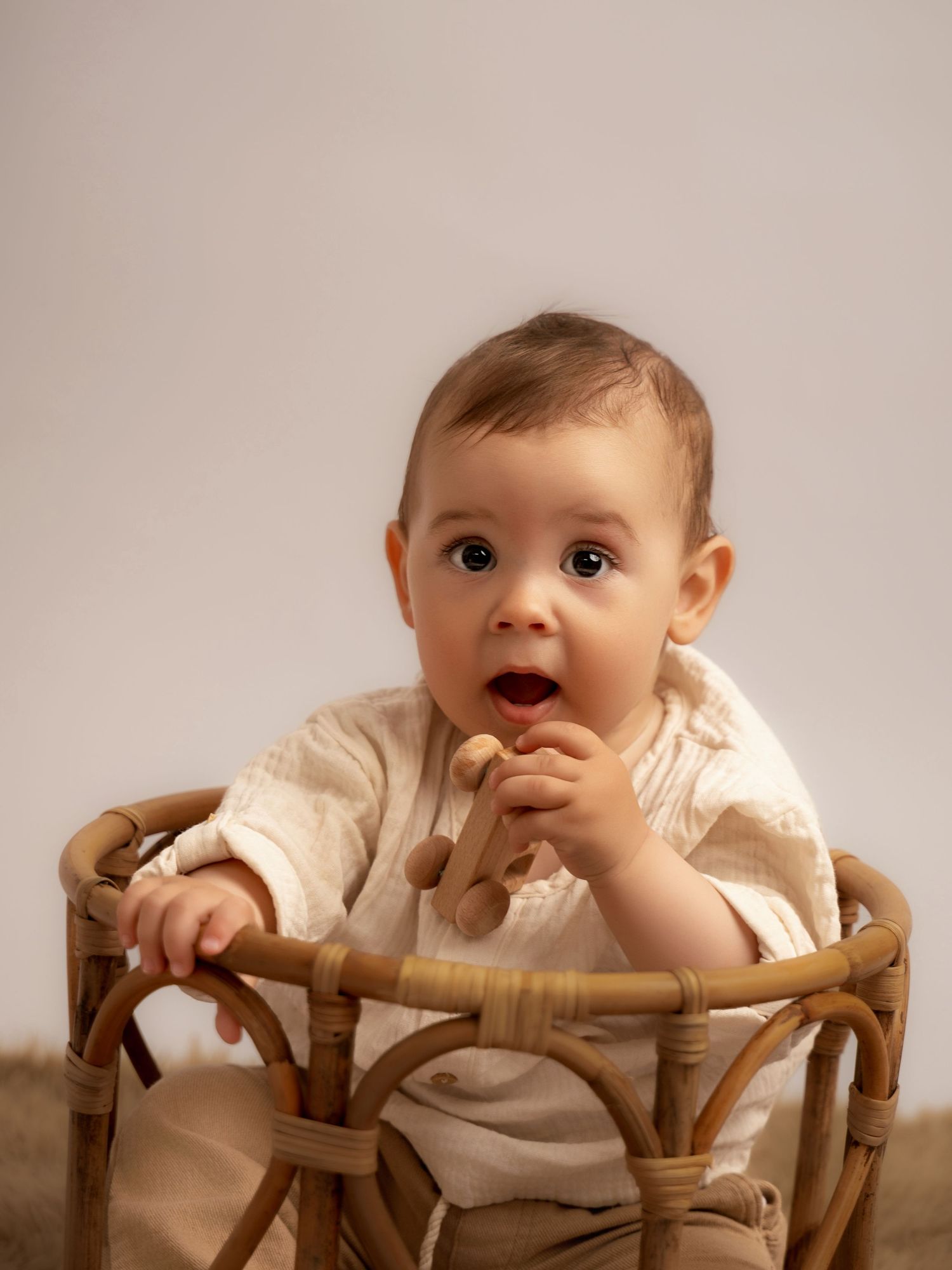 Adorable baby sitting in a rattan chair, chewing a wooden toy, wearing a cream outfit against a neutral background.