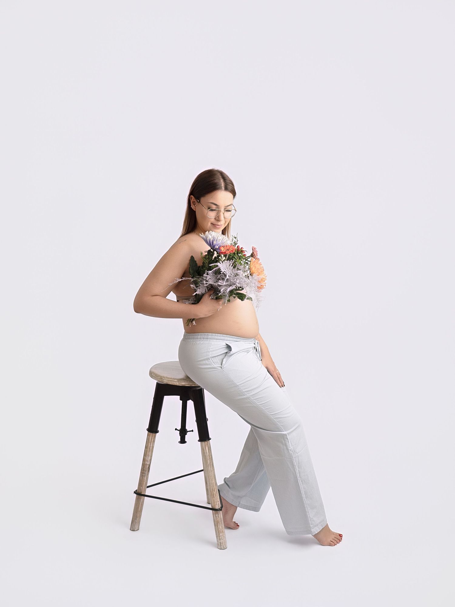 Pregnant woman sitting on stool holding colorful flowers, wearing light gray maternity pants on white background.