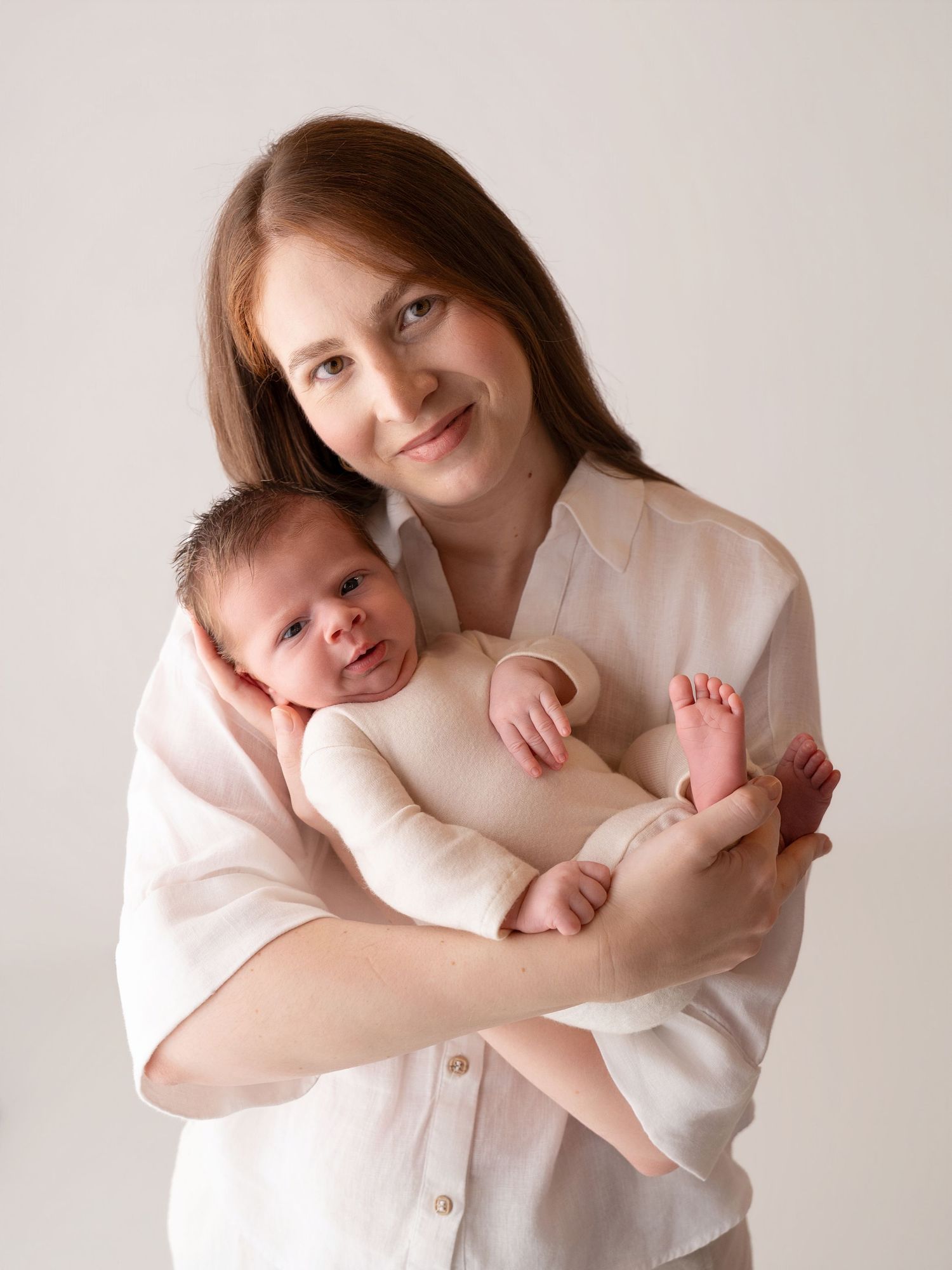 A smiling mother in a white shirt tenderly holds her newborn baby against a soft neutral background.