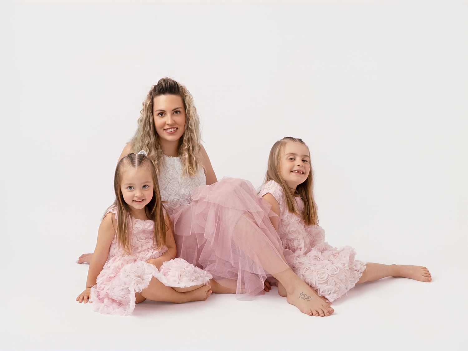 A mother and two young daughters pose together in matching pink dresses against a white background.