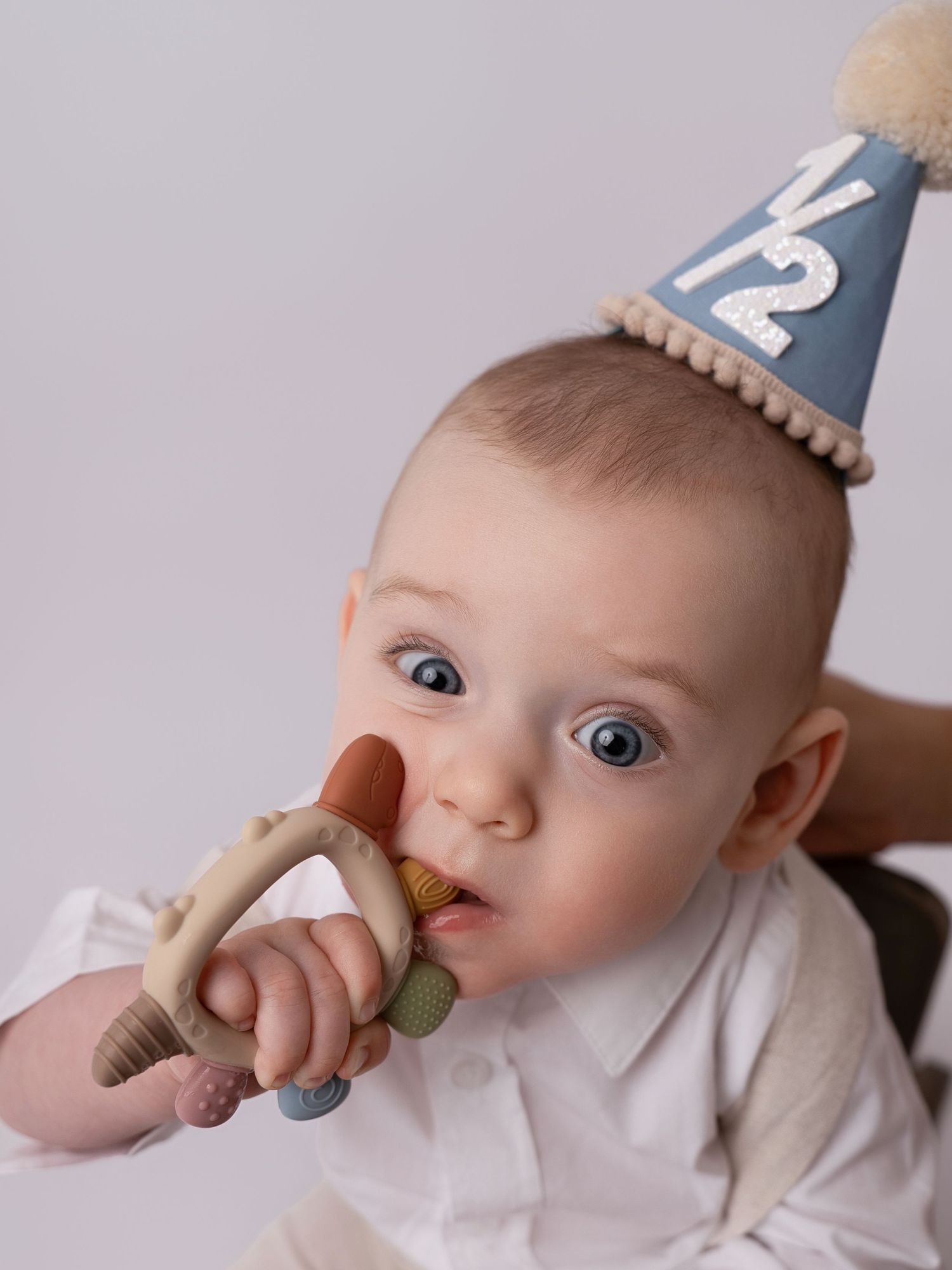 A blue-eyed baby wearing a blue half-birthday party hat chews on a wooden teething ring toy.