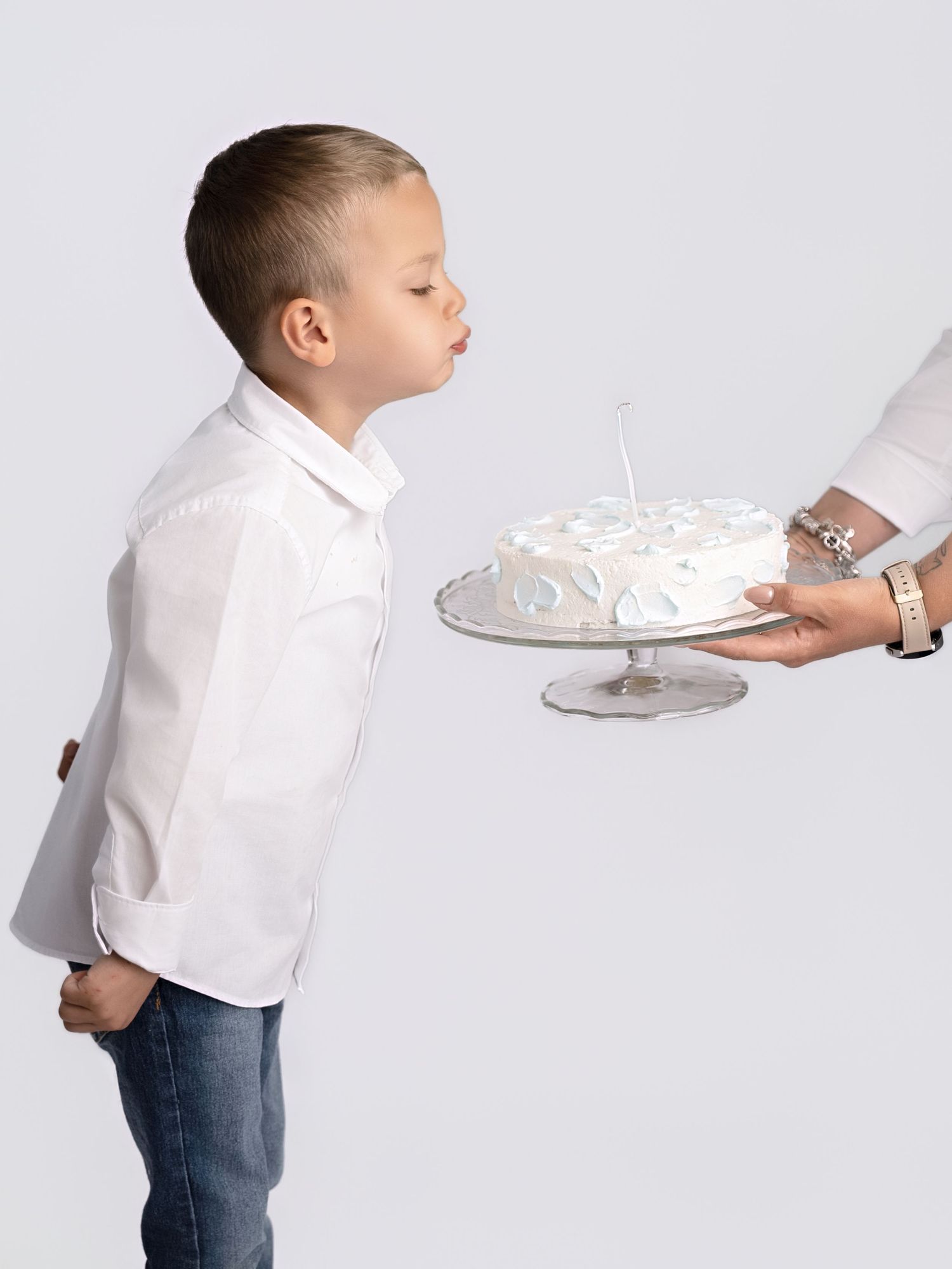 Young boy in white shirt blows out candle on white birthday cake held by adult.