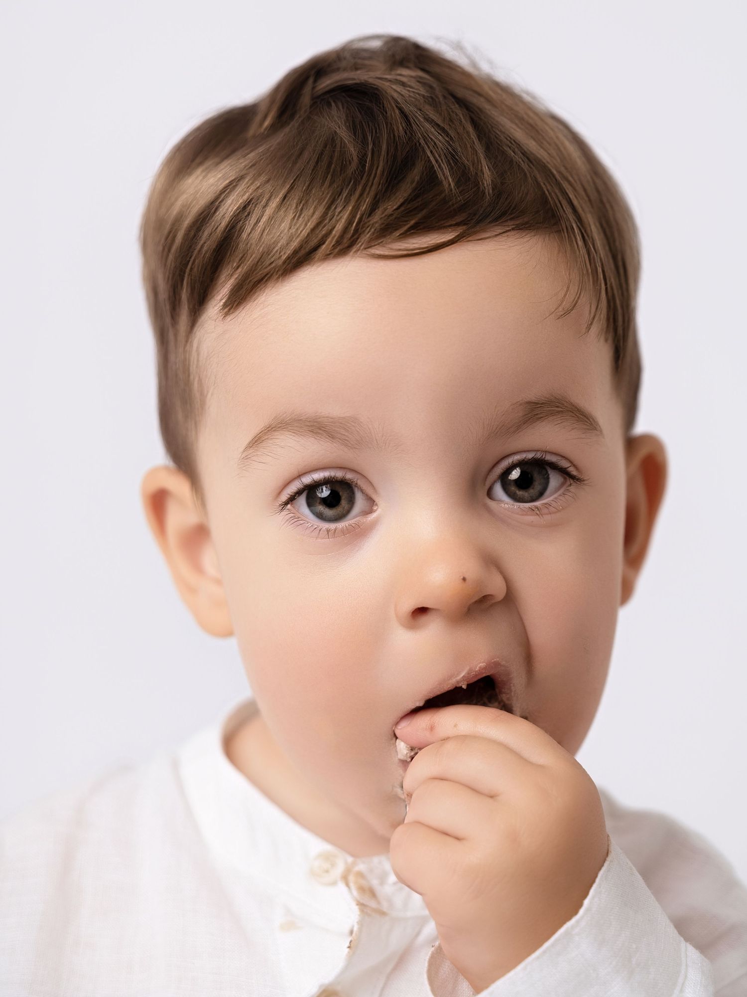 Adorable toddler boy with brown hair eating cake, wearing a white shirt against a light background.