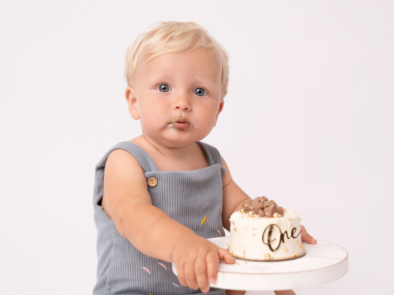 Blonde toddler in gray romper celebrating first birthday with white cake labeled One on white background.