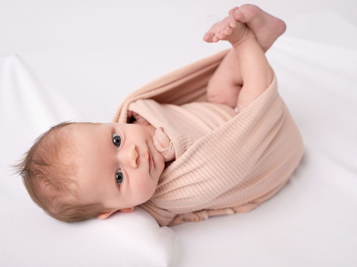 Adorable baby in a pink ribbed wrap lying on a white blanket, looking up with bright curious eyes.
