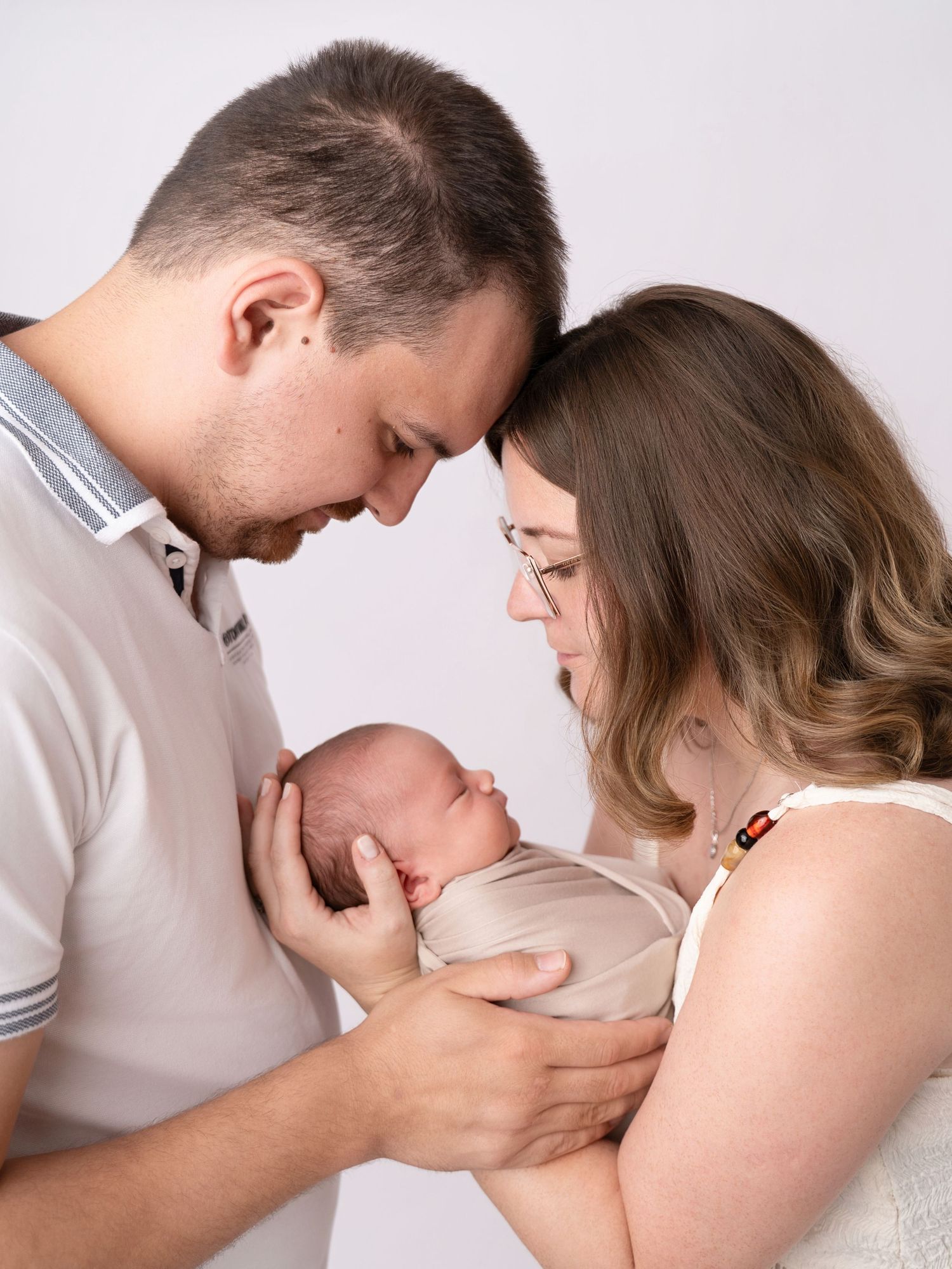 New parents tenderly hold their sleeping newborn baby together, foreheads touching in an intimate studio portrait.
