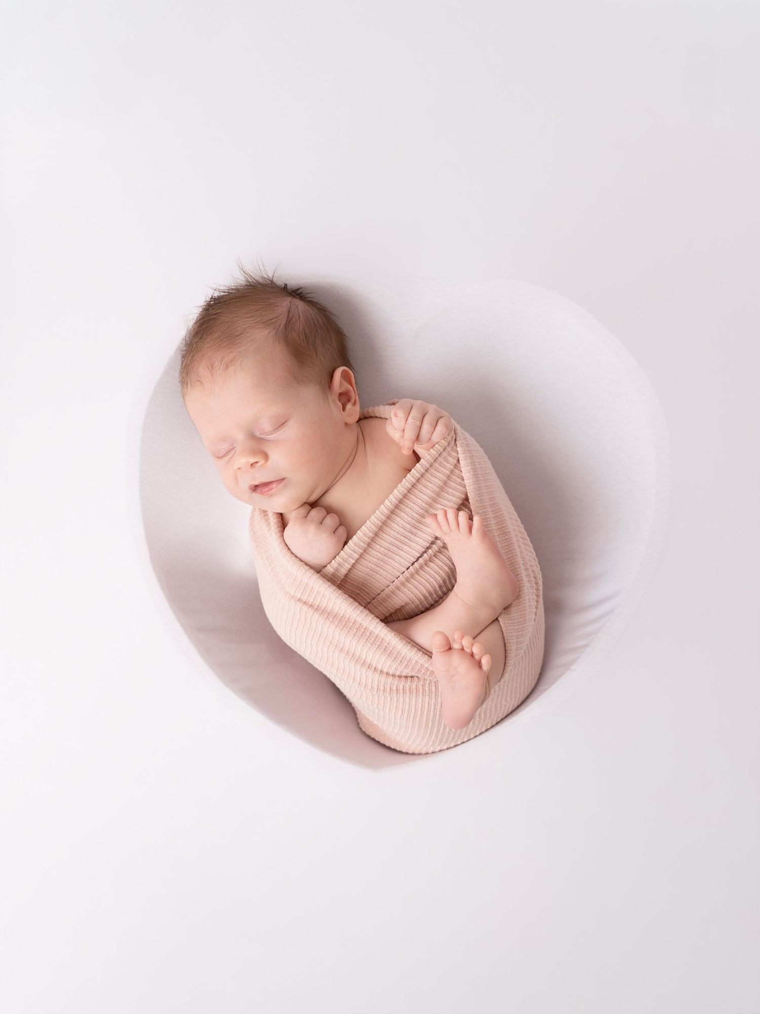 Newborn baby sleeping curled up in a round bowl, wearing a pink knit romper, on a white background.
