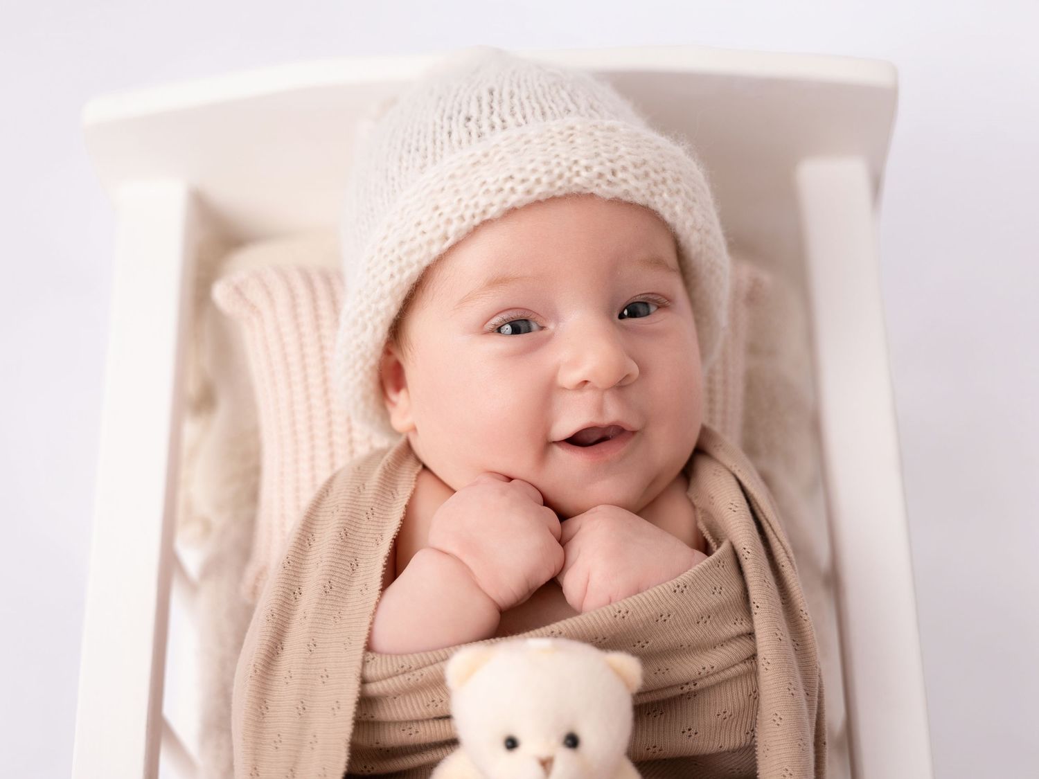 Smiling newborn baby in a knit hat wrapped in a beige blanket lying in a white crib with a teddy bear.