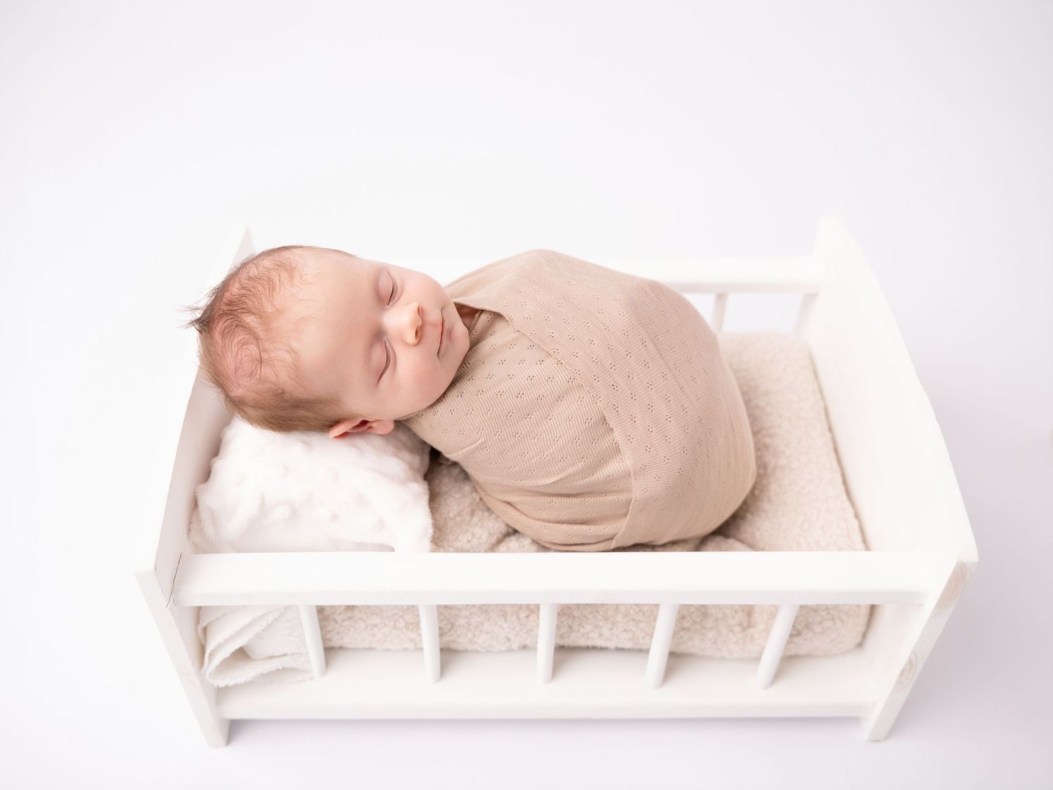 Sleeping newborn baby swaddled in beige wrap lying in a small white wooden cot on a white background.