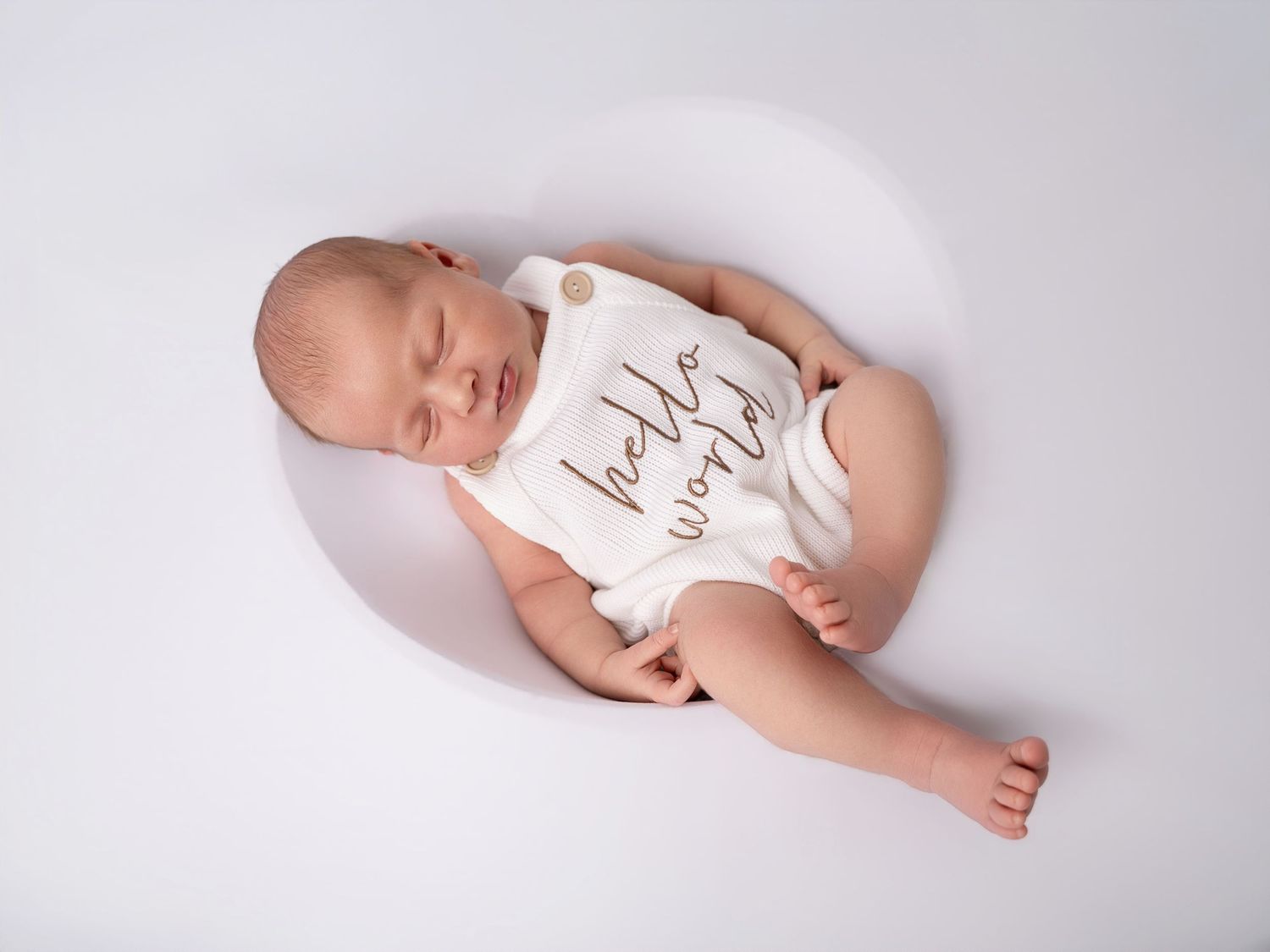 Sleeping newborn baby wearing a white 'hello world' onesie, photographed from above on a soft gray background.