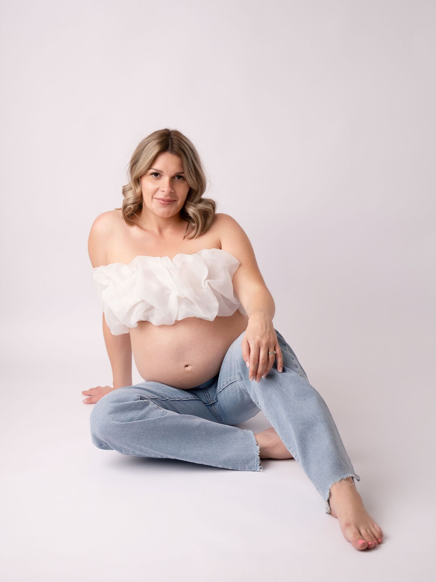 Pregnant woman in white ruffled crop top and jeans sitting on floor, showcasing baby bump in maternity photo shoot.
