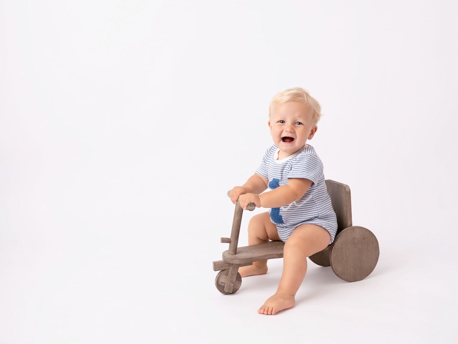 Happy baby boy in blue romper sitting on a wooden toy tricycle against a white background.