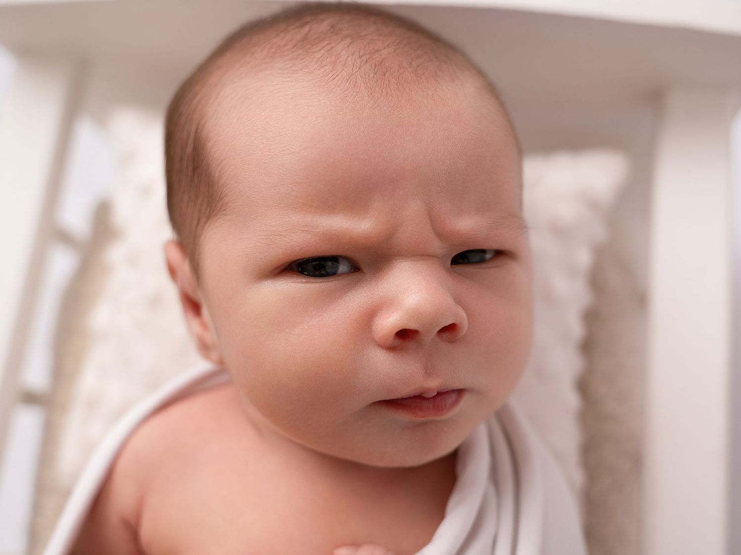 Newborn baby with a furrowed brow and skeptical expression wrapped in white swaddle blanket in a crib.