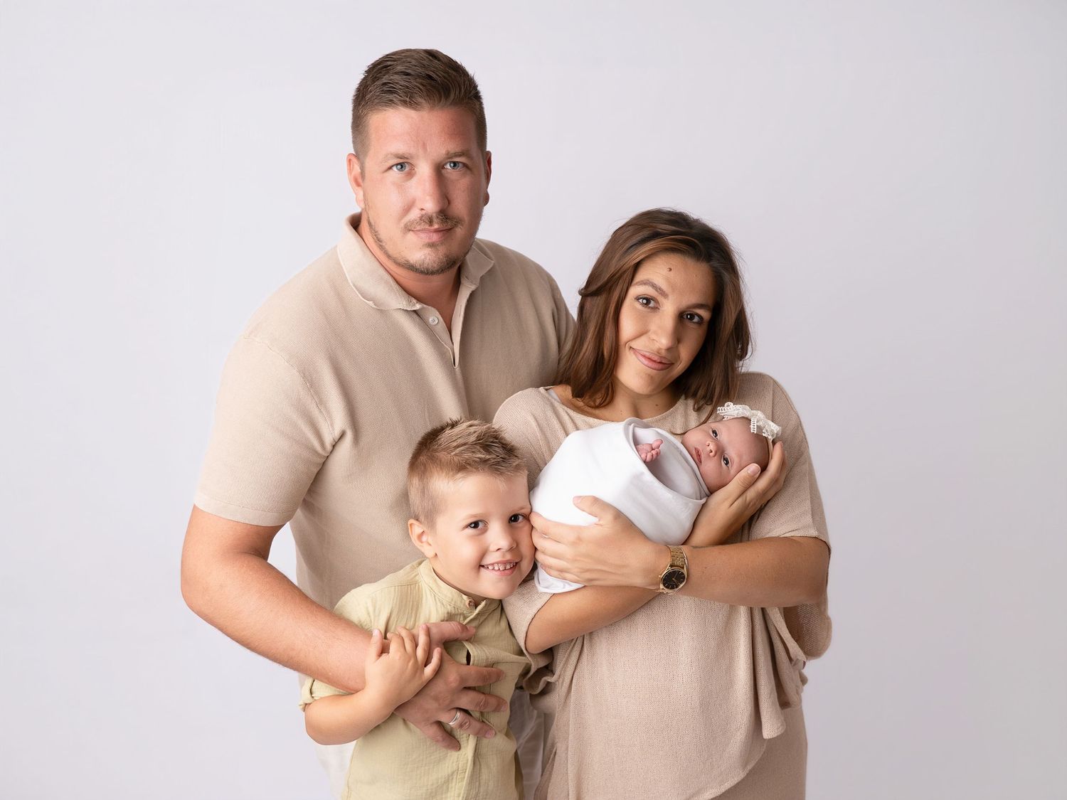 Family portrait with father, mother, toddler son, and swaddled newborn in matching beige outfits.