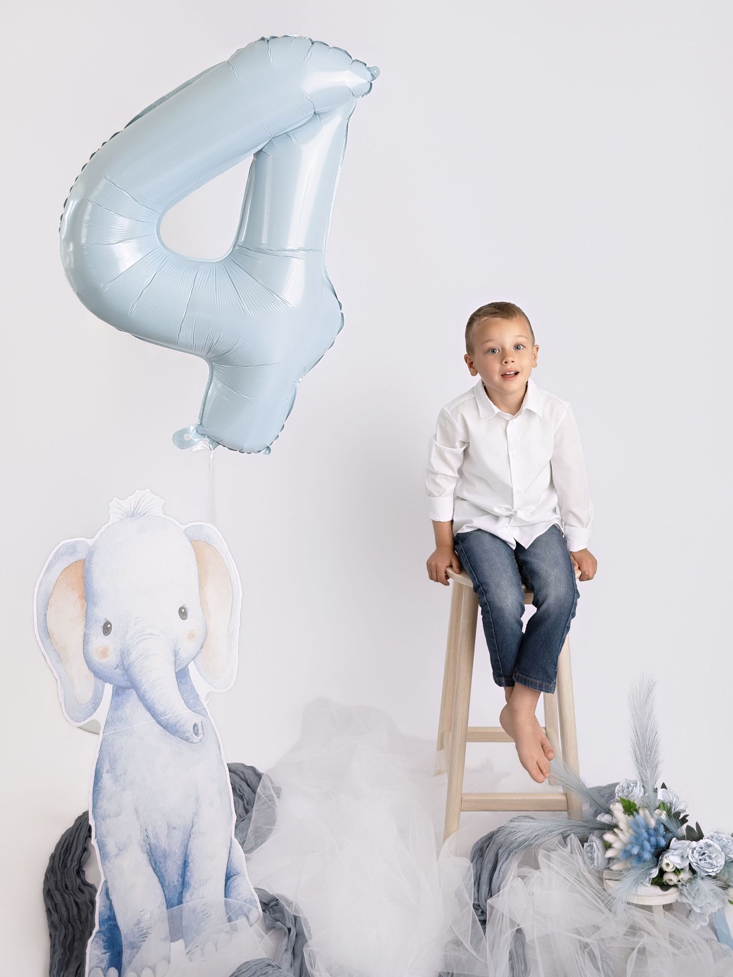 Boy in white shirt sits on stool beside number 4 balloon and elephant cutout for 4th birthday.