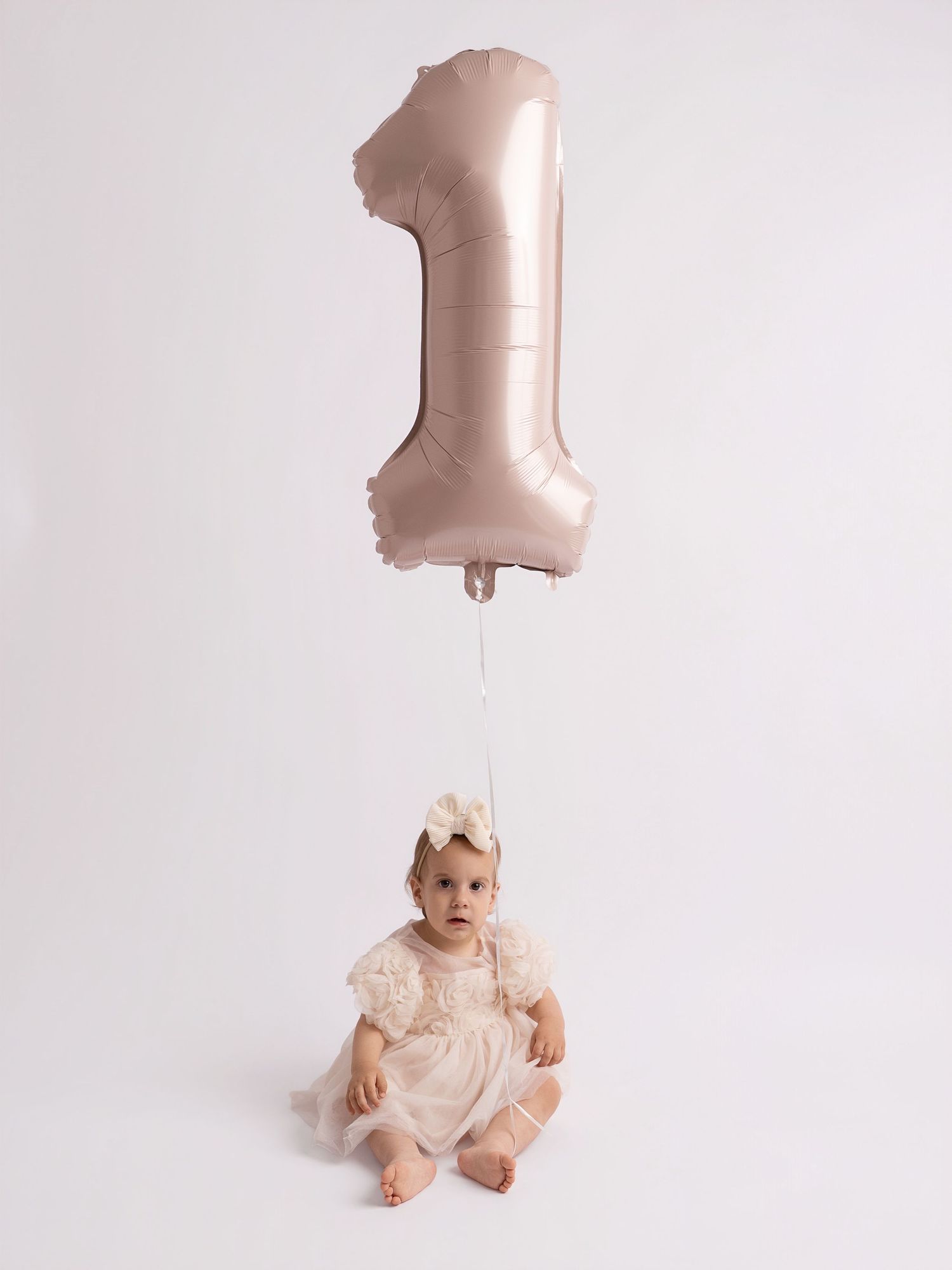 Baby girl in white lace dress sits beneath a rose gold number 1 balloon celebrating her first birthday.