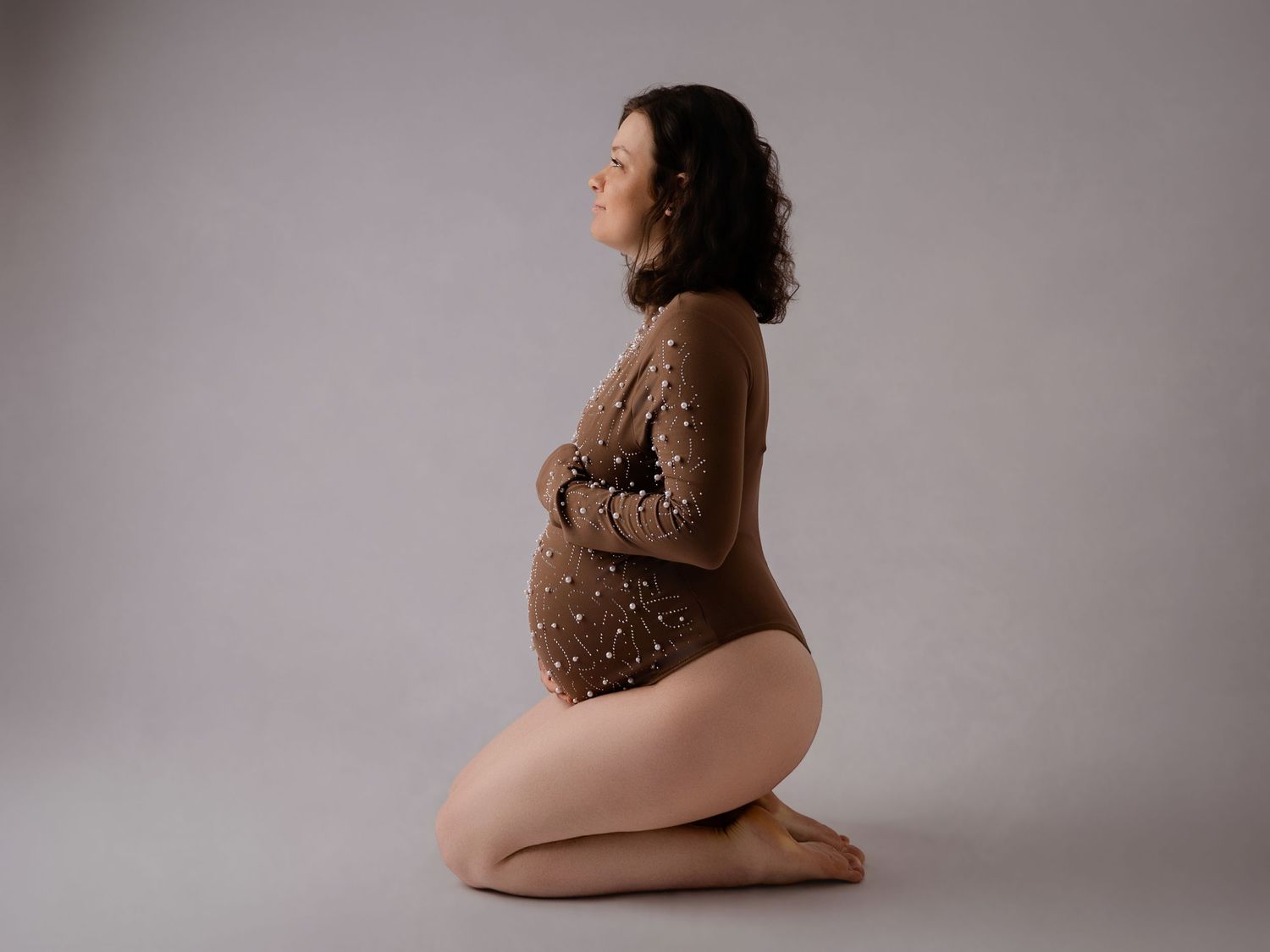 Side profile of pregnant woman kneeling in brown bodysuit showcasing her baby bump.