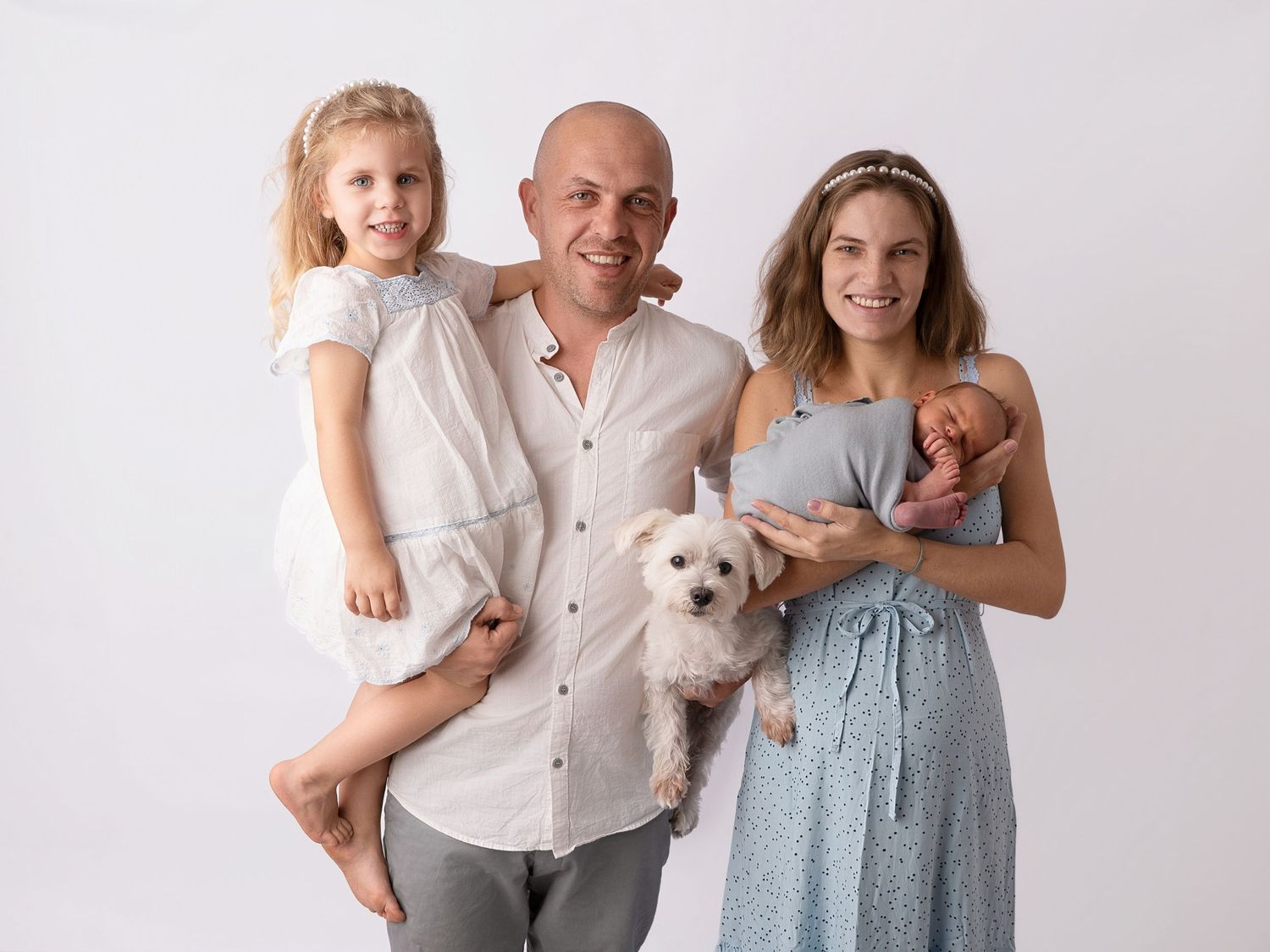 Family portrait with bald man holding toddler girl and small white dog, woman in blue dress holding newborn, white background.