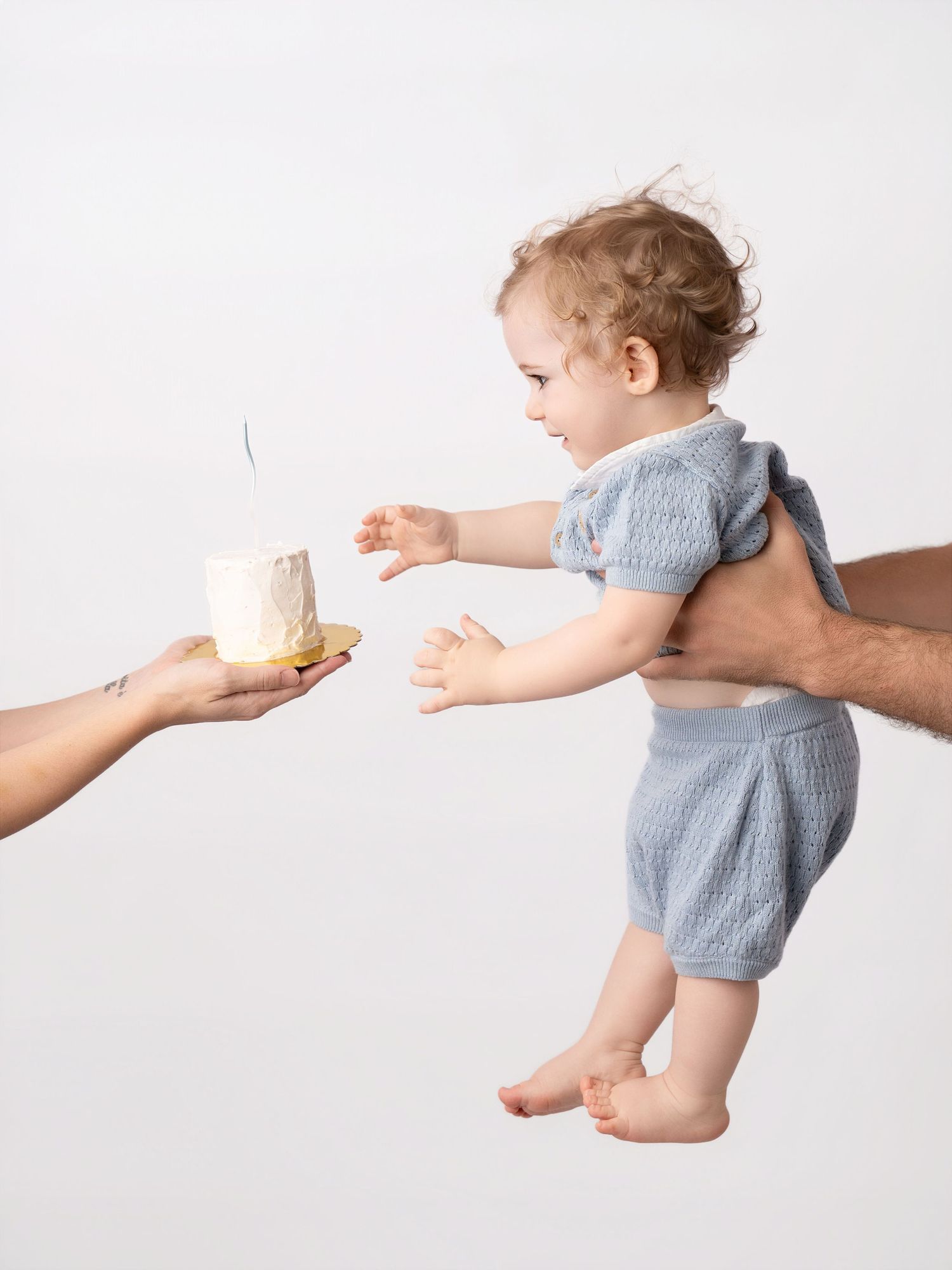 Adult holding baby reaching for small birthday smash cake on white background.
