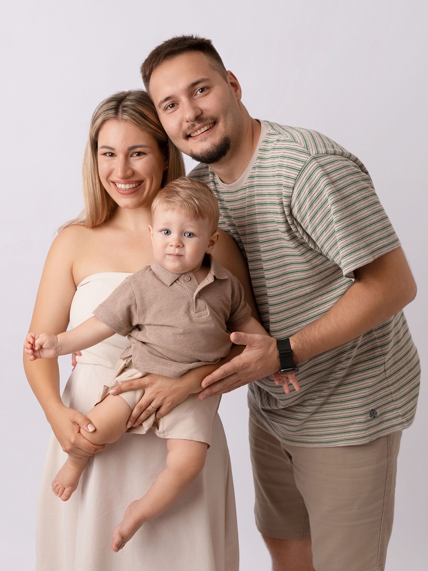 A smiling young family with a blonde mother, father in striped shirt, and baby boy posing together in a studio.