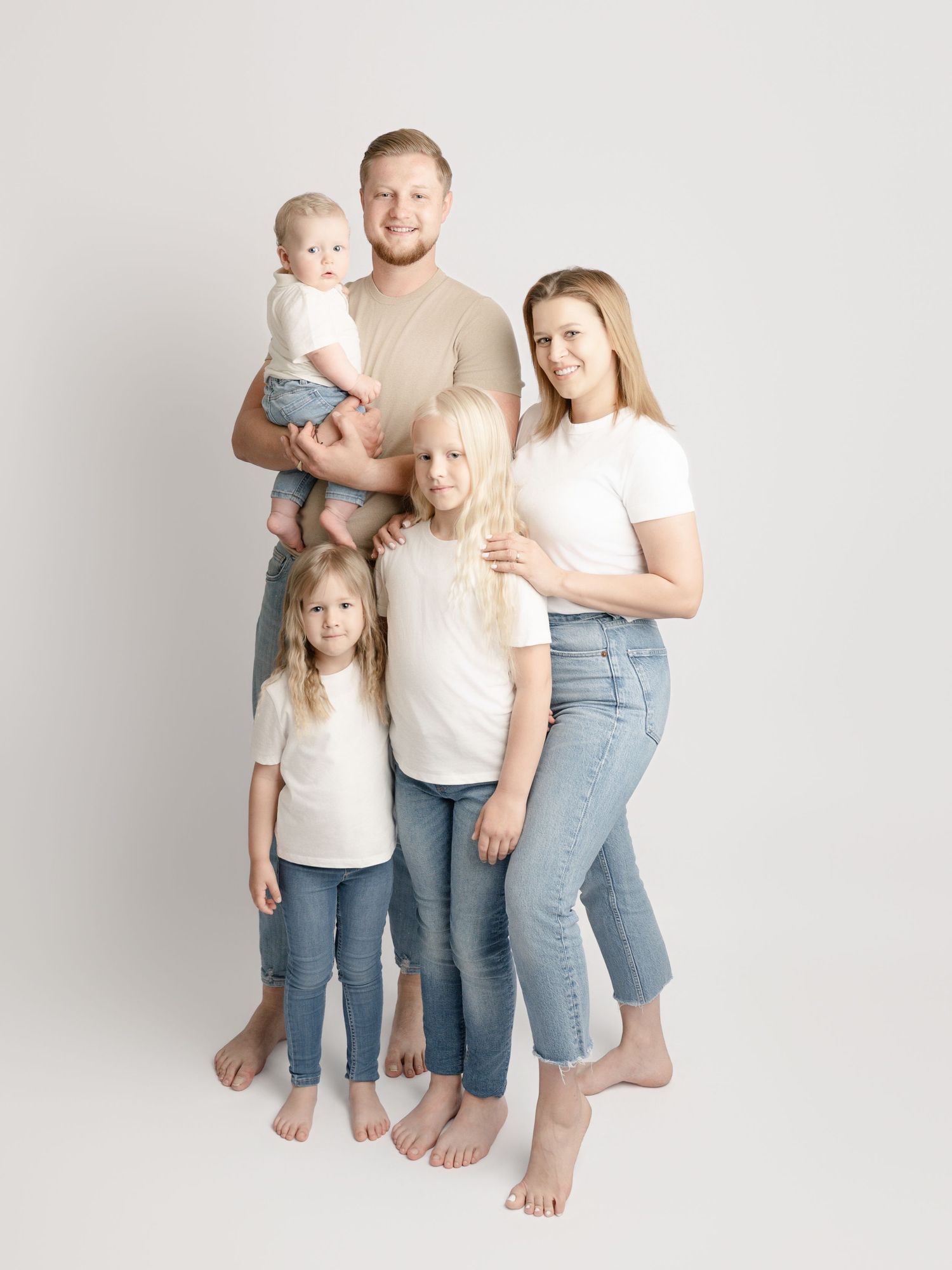 A family of five poses together in matching white tops and jeans, including three children and a baby, in a bright studio.