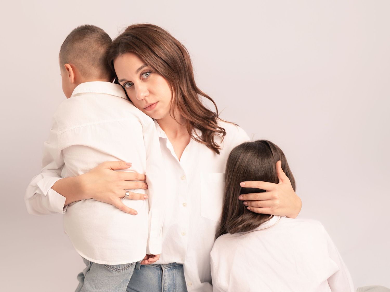 Mother in white shirt embracing two children in matching white outfits in bright studio portrait.