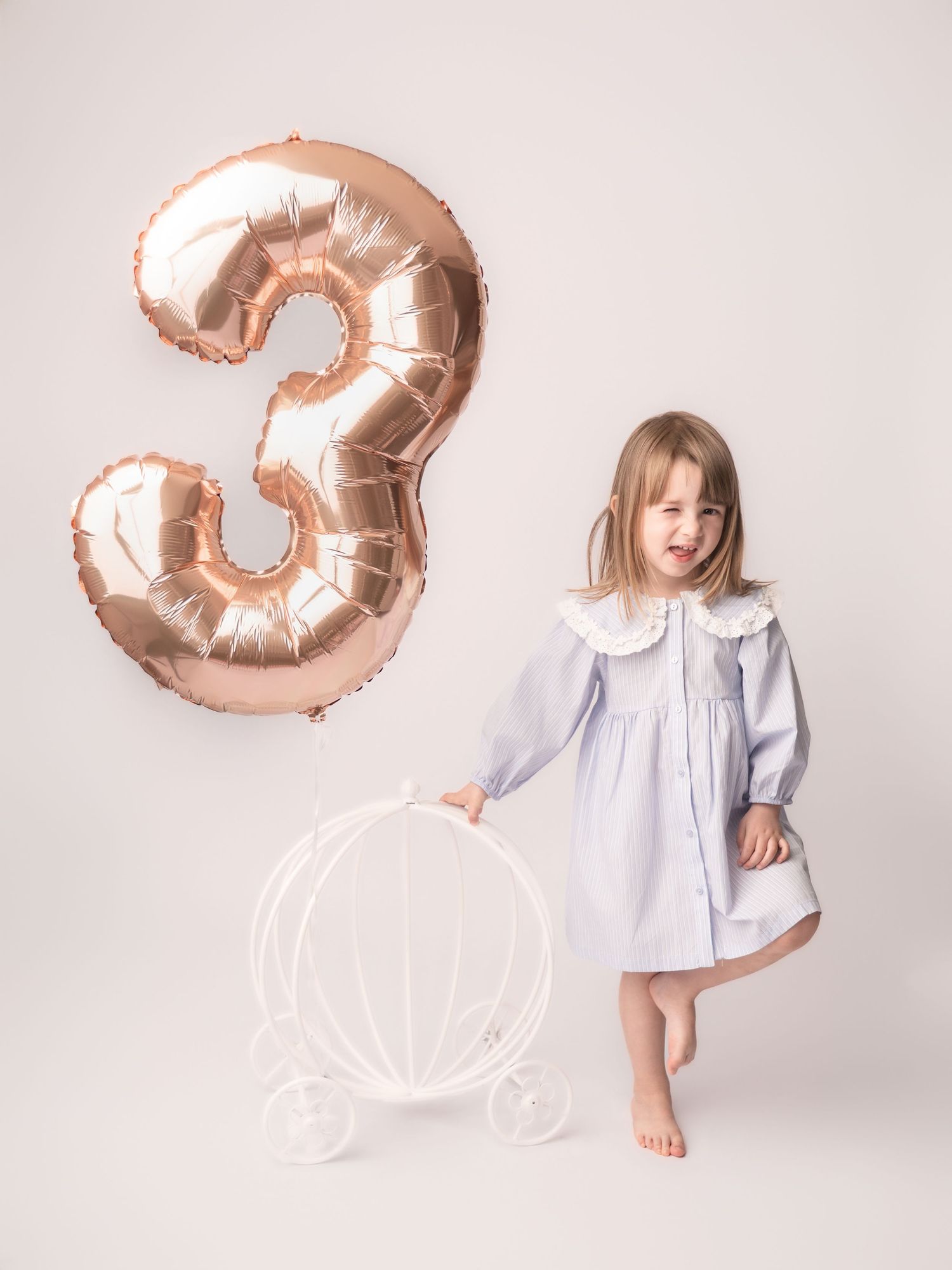 A smiling little girl in a blue dress stands next to a rose gold number 3 balloon and white carriage prop.