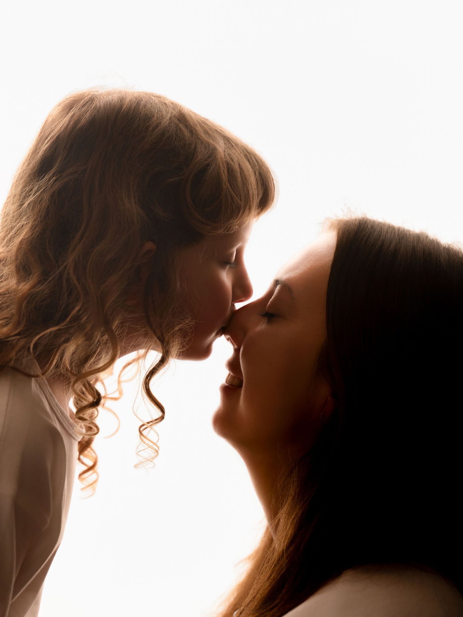 A mother and young daughter share a tender nose-to-nose moment in a warm backlit portrait.