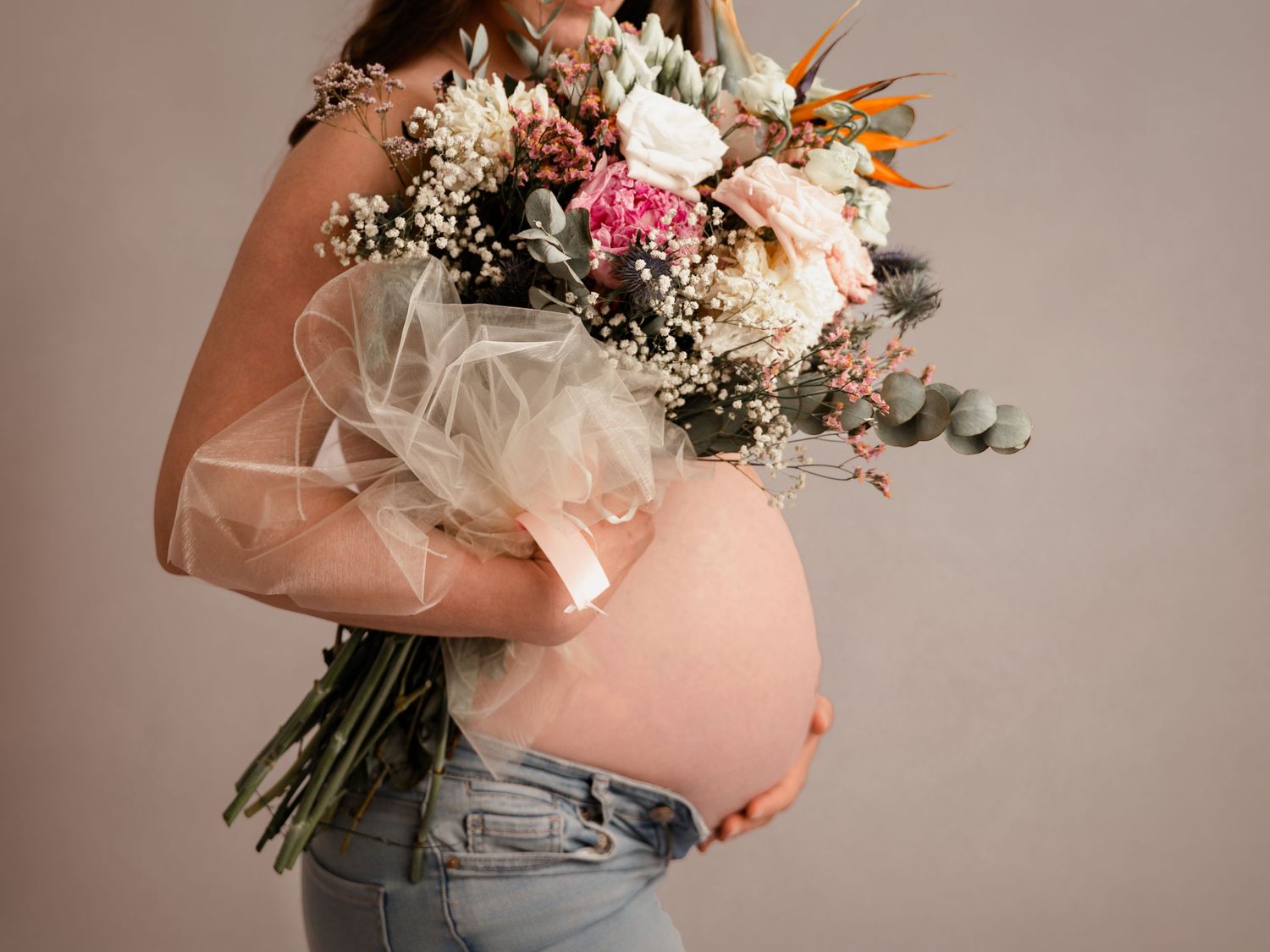 Pregnant woman in jeans holding a beautiful bouquet of mixed flowers against a neutral background.