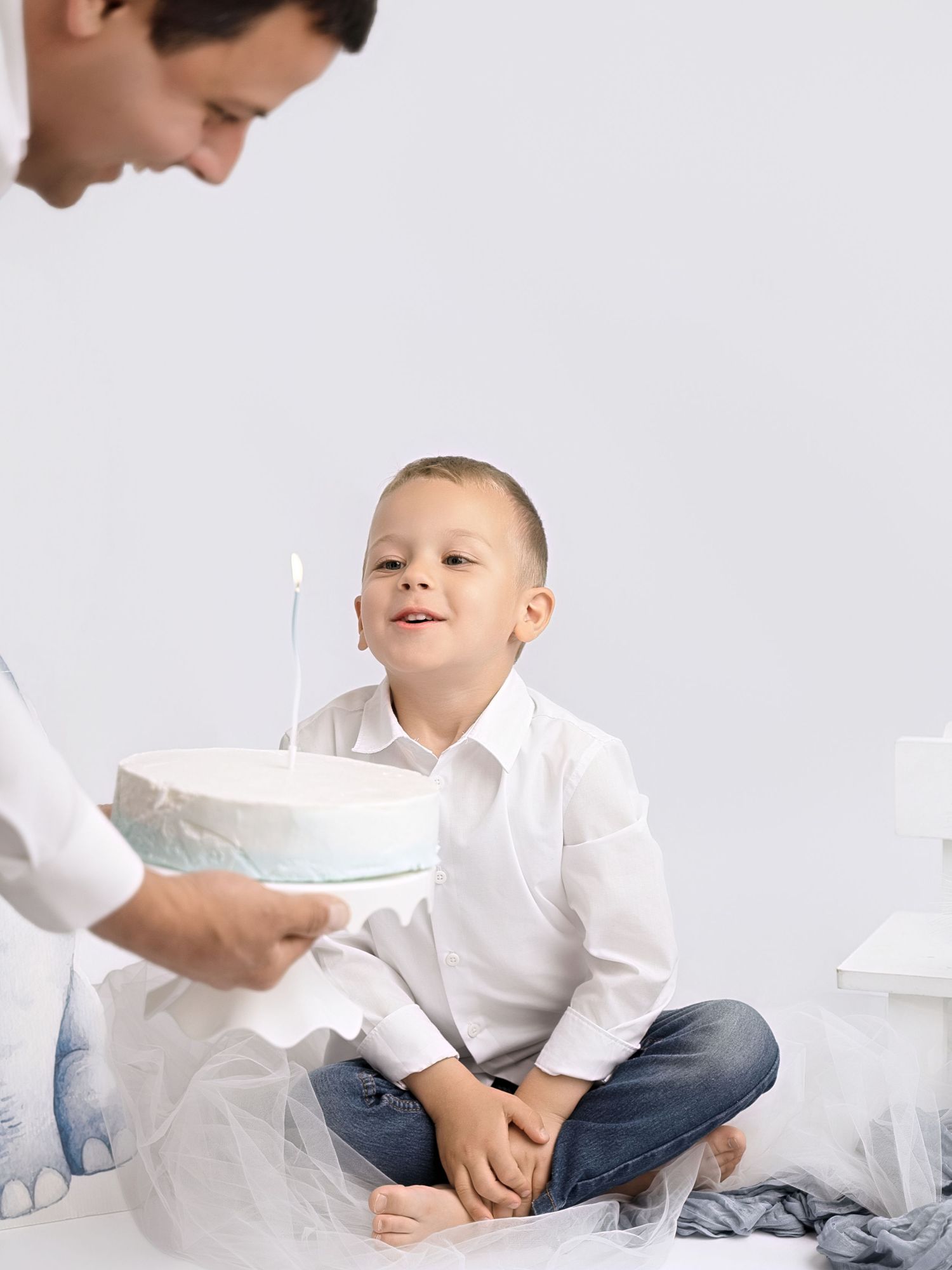 A young boy smiles at a birthday cake with a candle held by an adult in a bright white setting.
