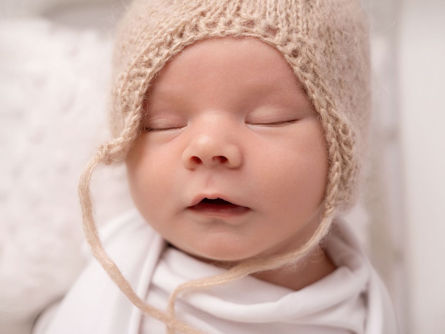 Peaceful newborn baby sleeping in a cozy knit beige bonnet hat, wrapped in white swaddle cloth.