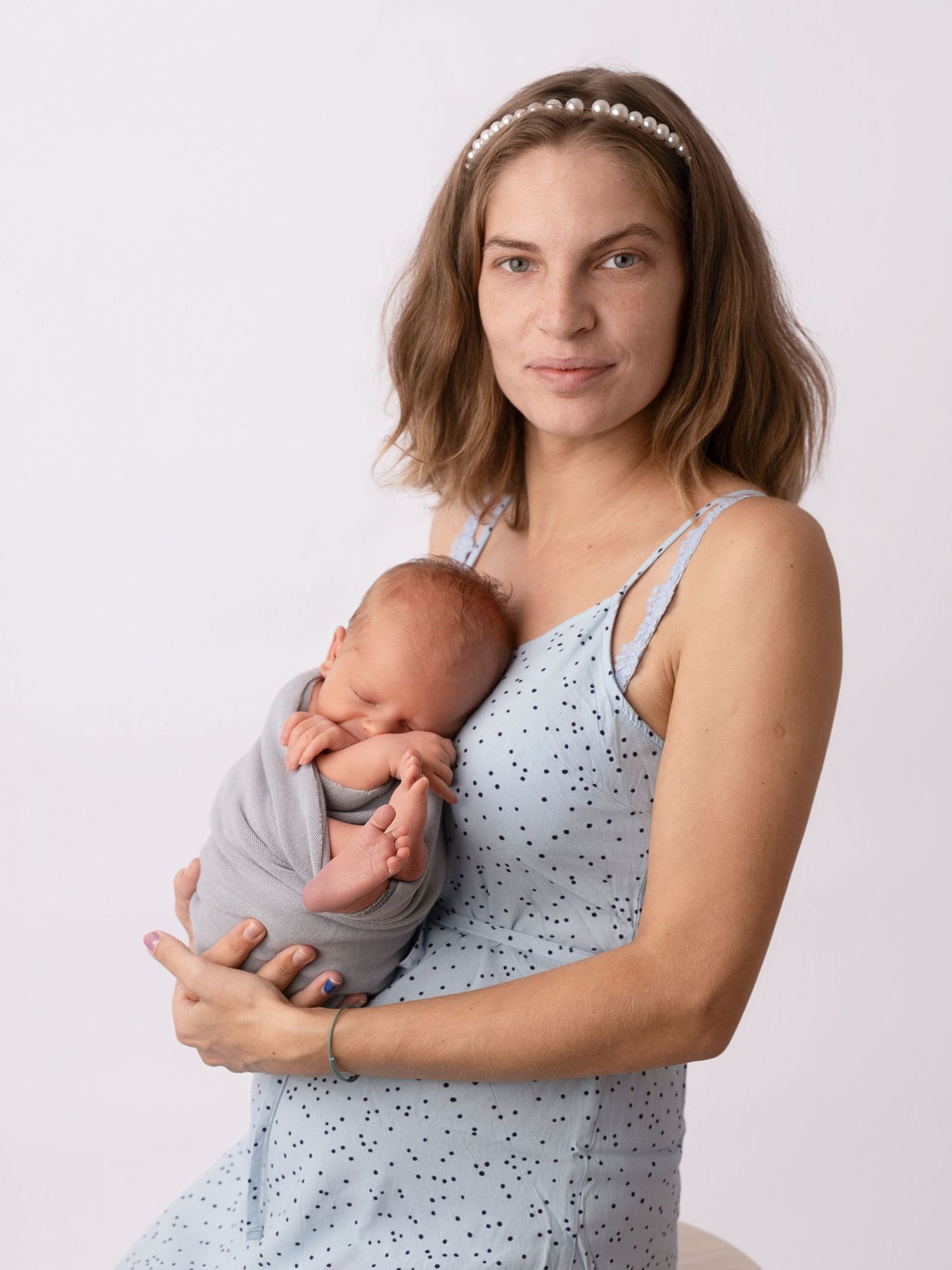 Young mother in blue dress holds sleeping newborn baby wrapped in gray swaddle, studio portrait.